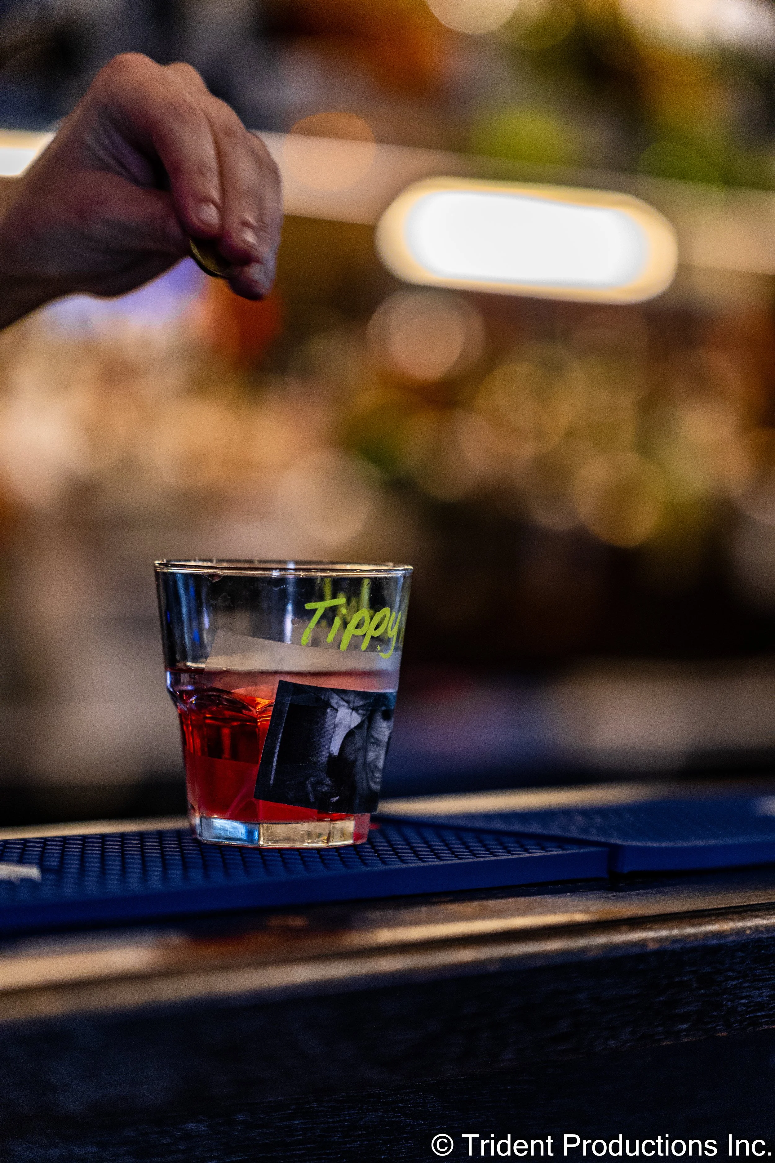 Close-up of a hand sprinkling something into a glass of red drink labeled 'Tippy' on a bar counter, with blurred colorful background lights.