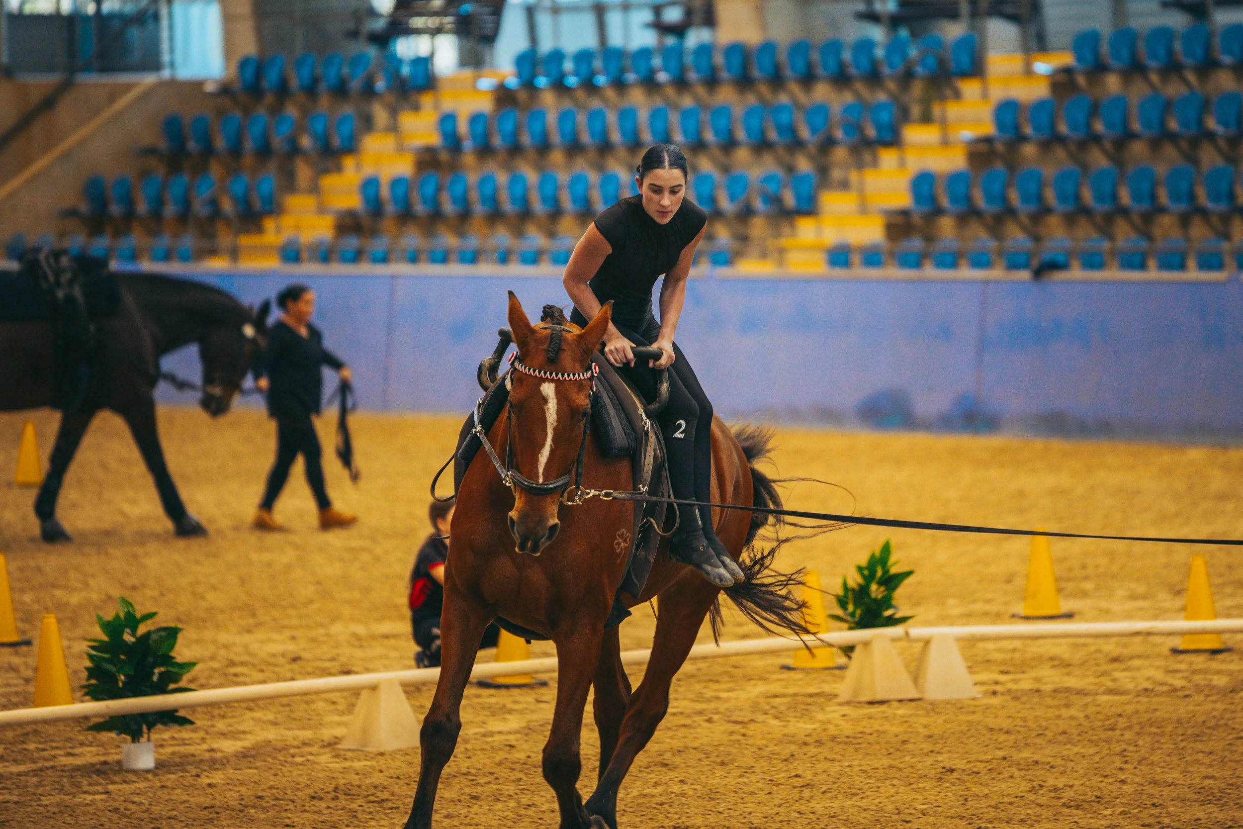 A woman riding a brown horse in an indoor arena with blue seats and yellow cones, with another woman and horse in the background.