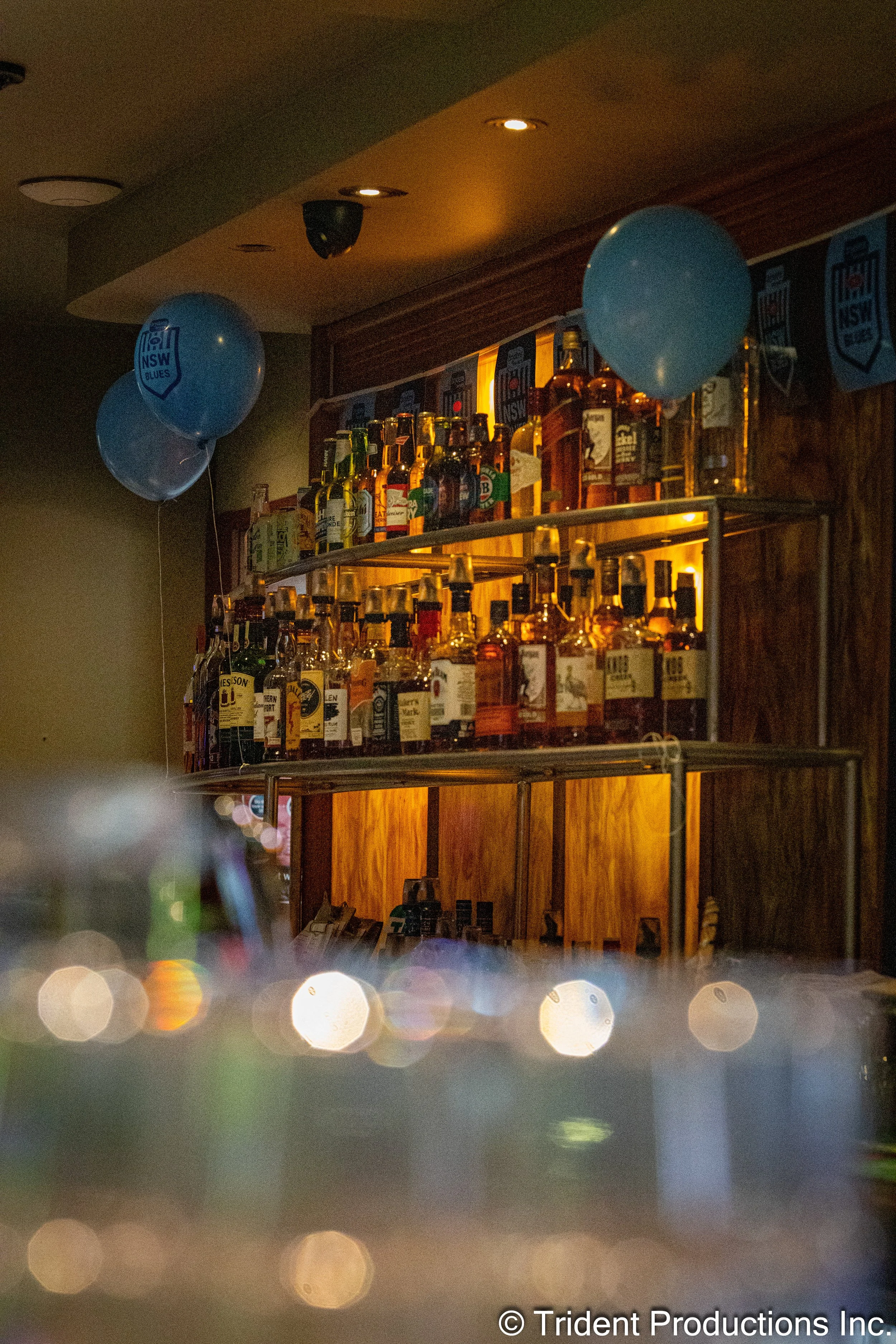 A bar shelf with various bottles of alcohol and two blue balloons with NSWR Blues logos hanging nearby.