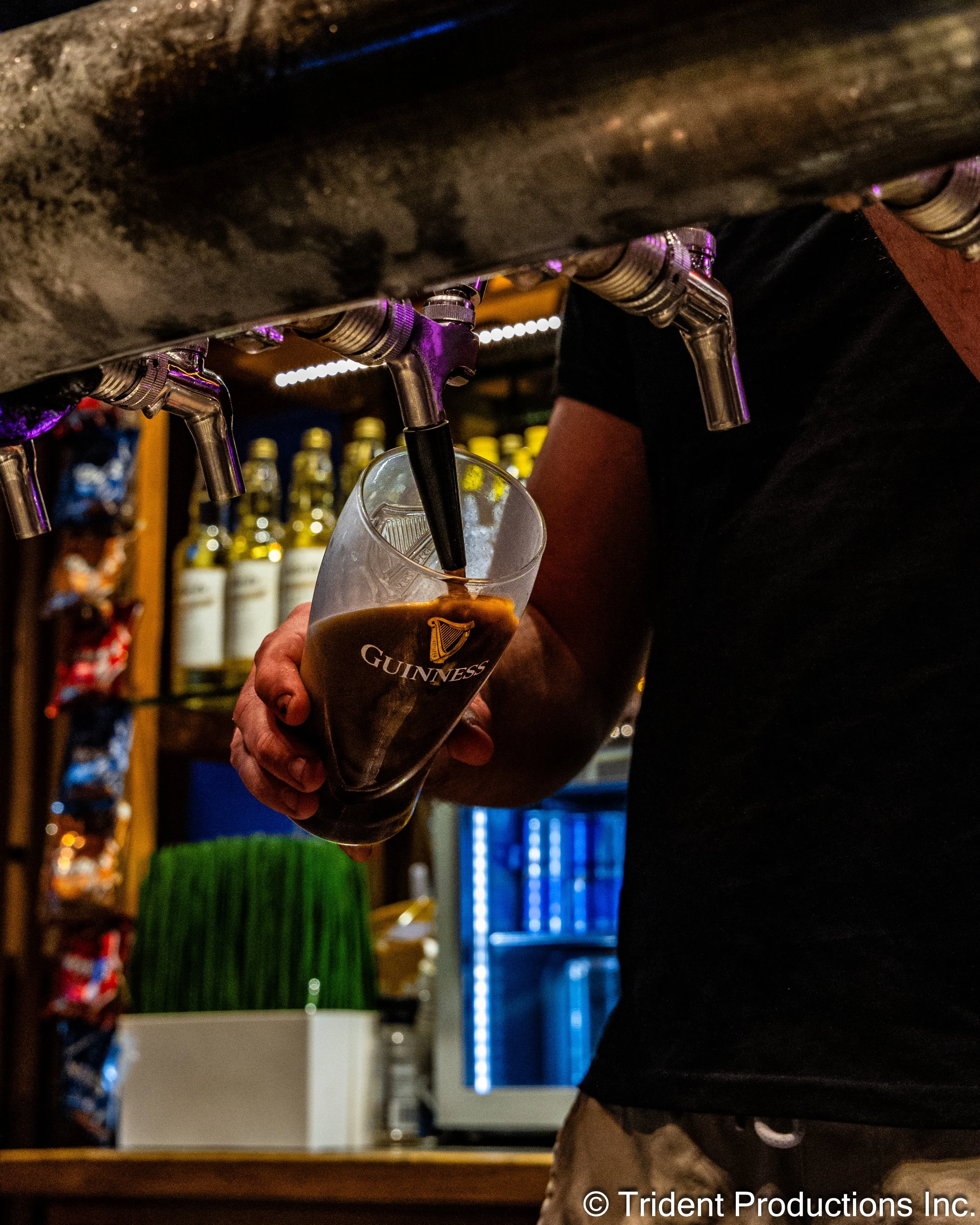 A bartender fills a Guinness beer glass from a tap at a bar, with bottles on shelves in the background.