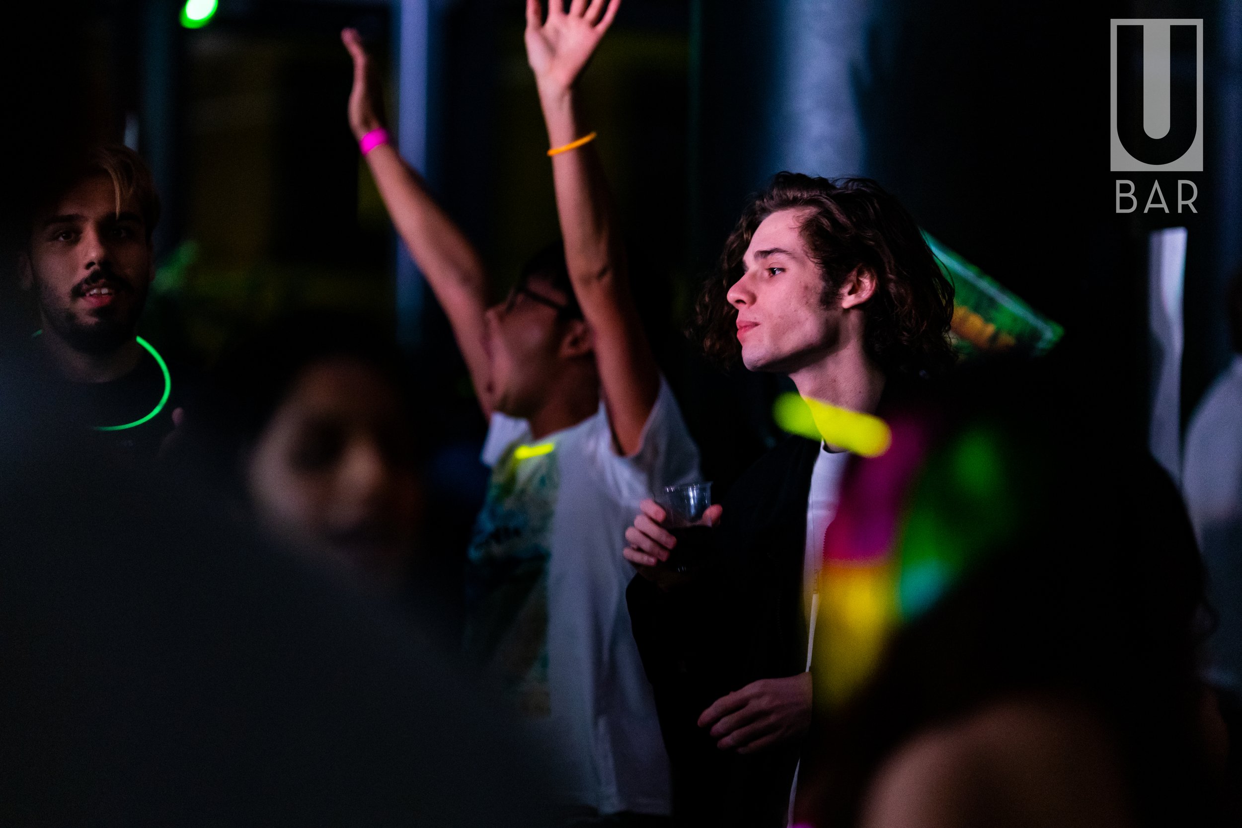 People dancing and socializing at a bar with colorful glow sticks, under dim lighting with a neon sign that reads 'U BAR'.
