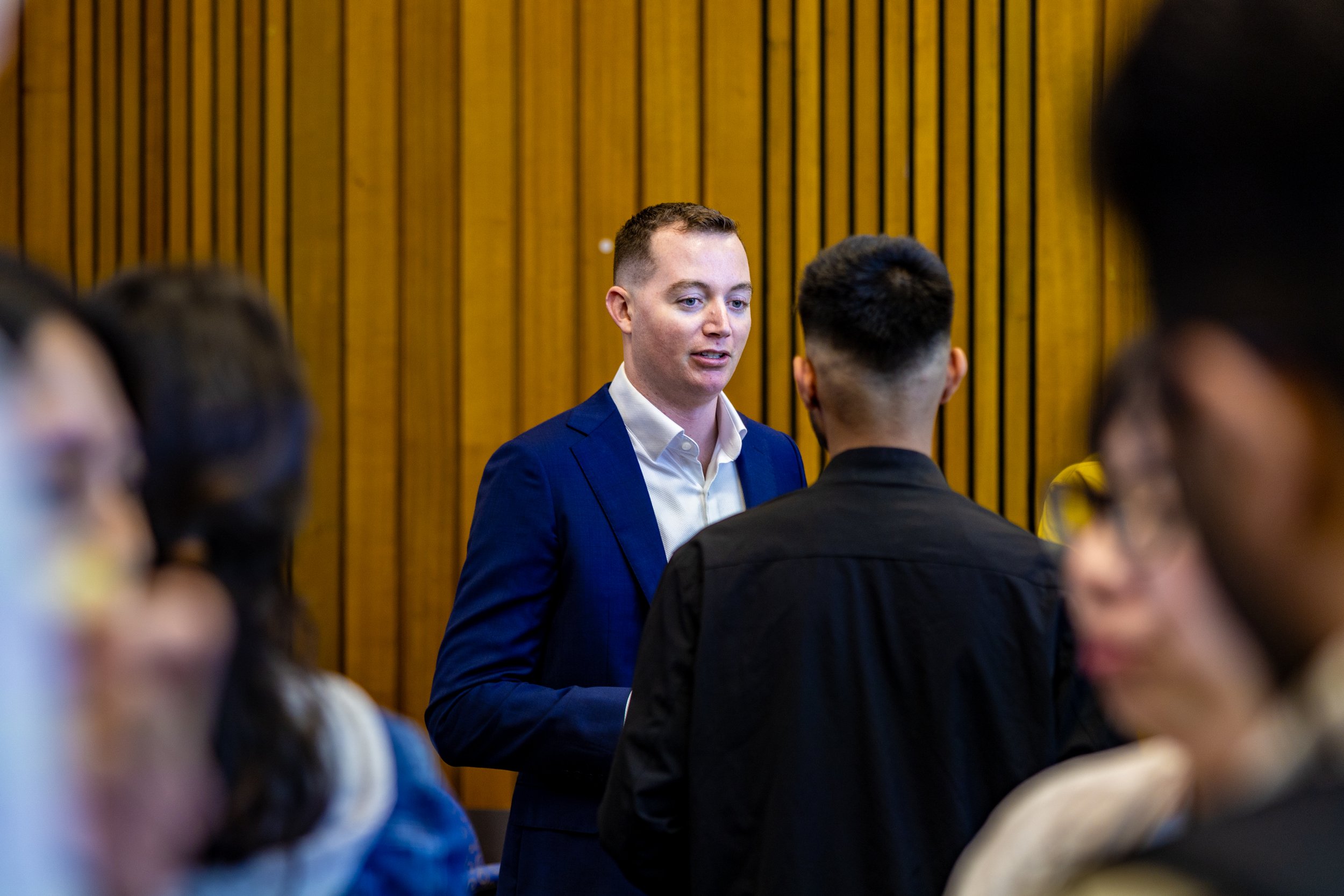 A man in a blue suit speaking to a group of people in a room with wooden panel walls.