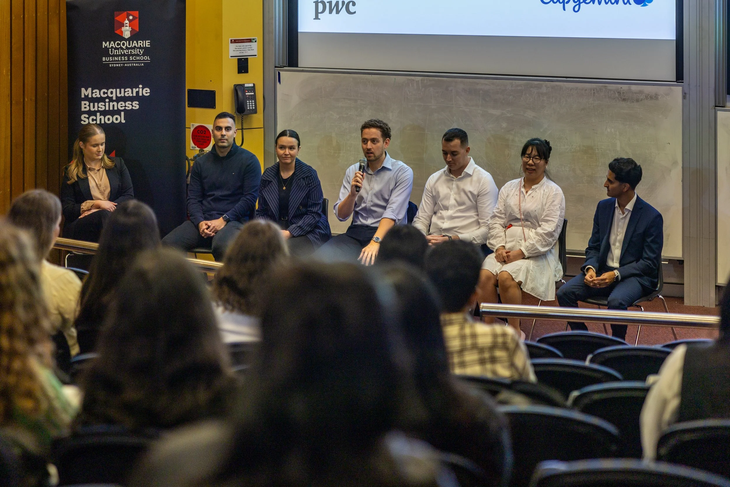 Panel discussion with seven people sitting on stage in front of an audience, at Macquarie University Business School in Sydney, Australia.