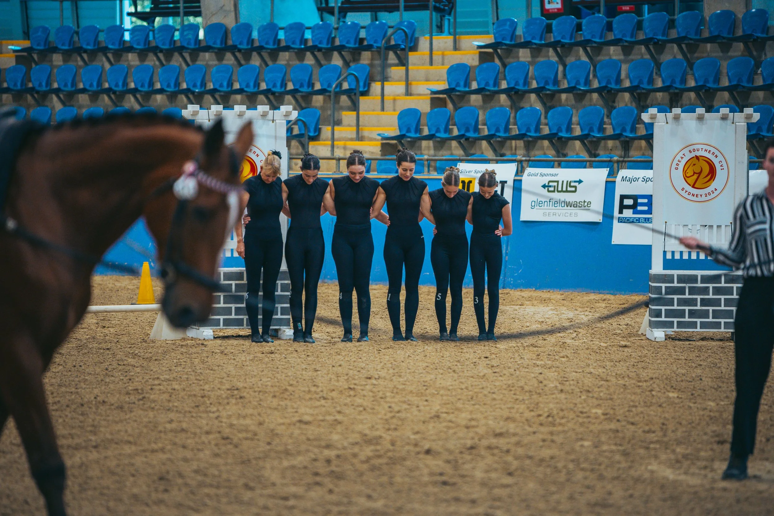 Six female equestrians in navy uniforms bowing in an indoor arena with blue seats, a horse in foreground, and racing barricades and banners in the background.