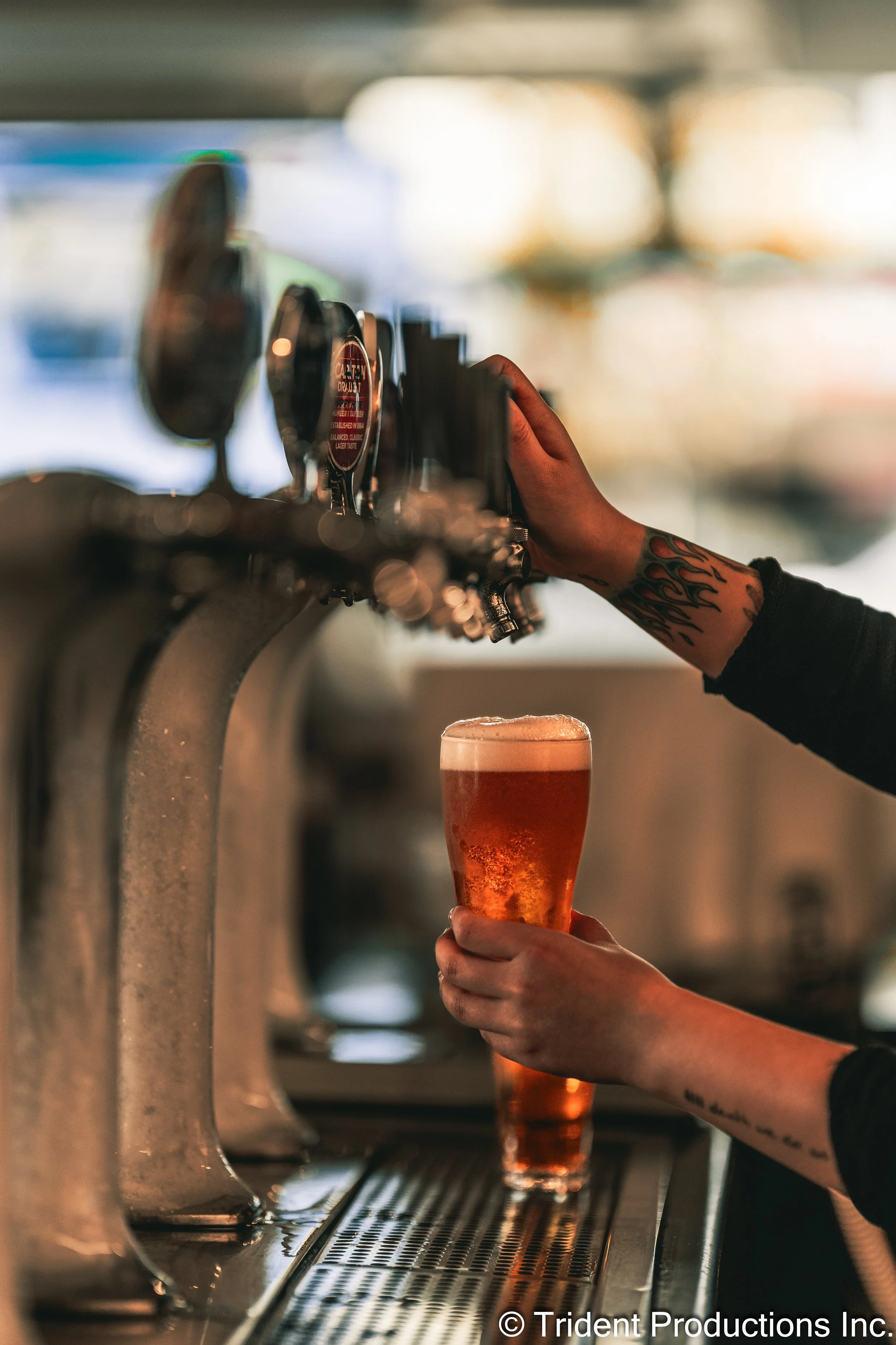 A person with a tattoo on their arm is pouring beer from a tap into a glass in a bar or brewery setting.