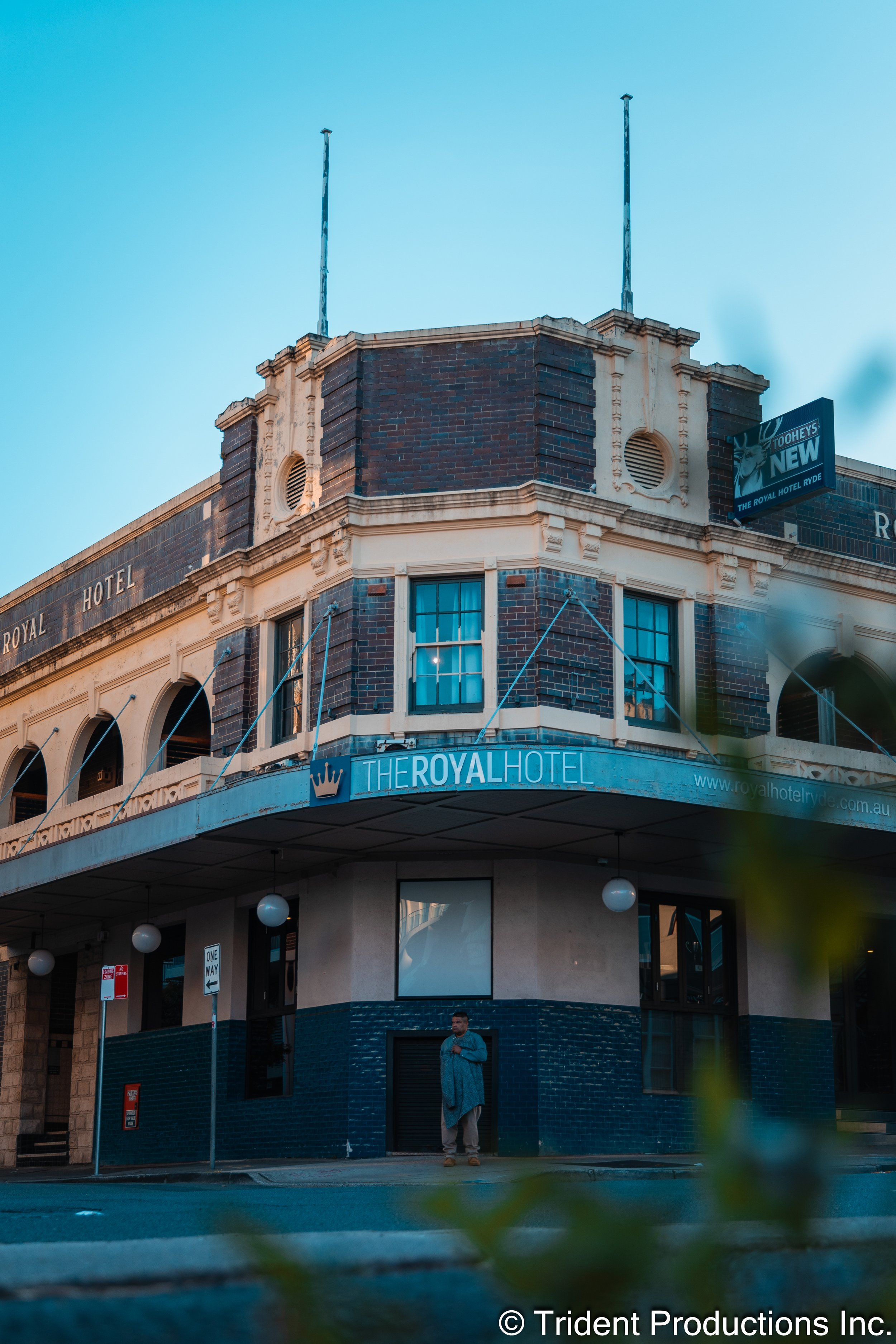 The Royal Hotel, a corner building with brick and beige architecture, has signs indicating its name and a billboard advertising its new status, with a man standing on the sidewalk below.