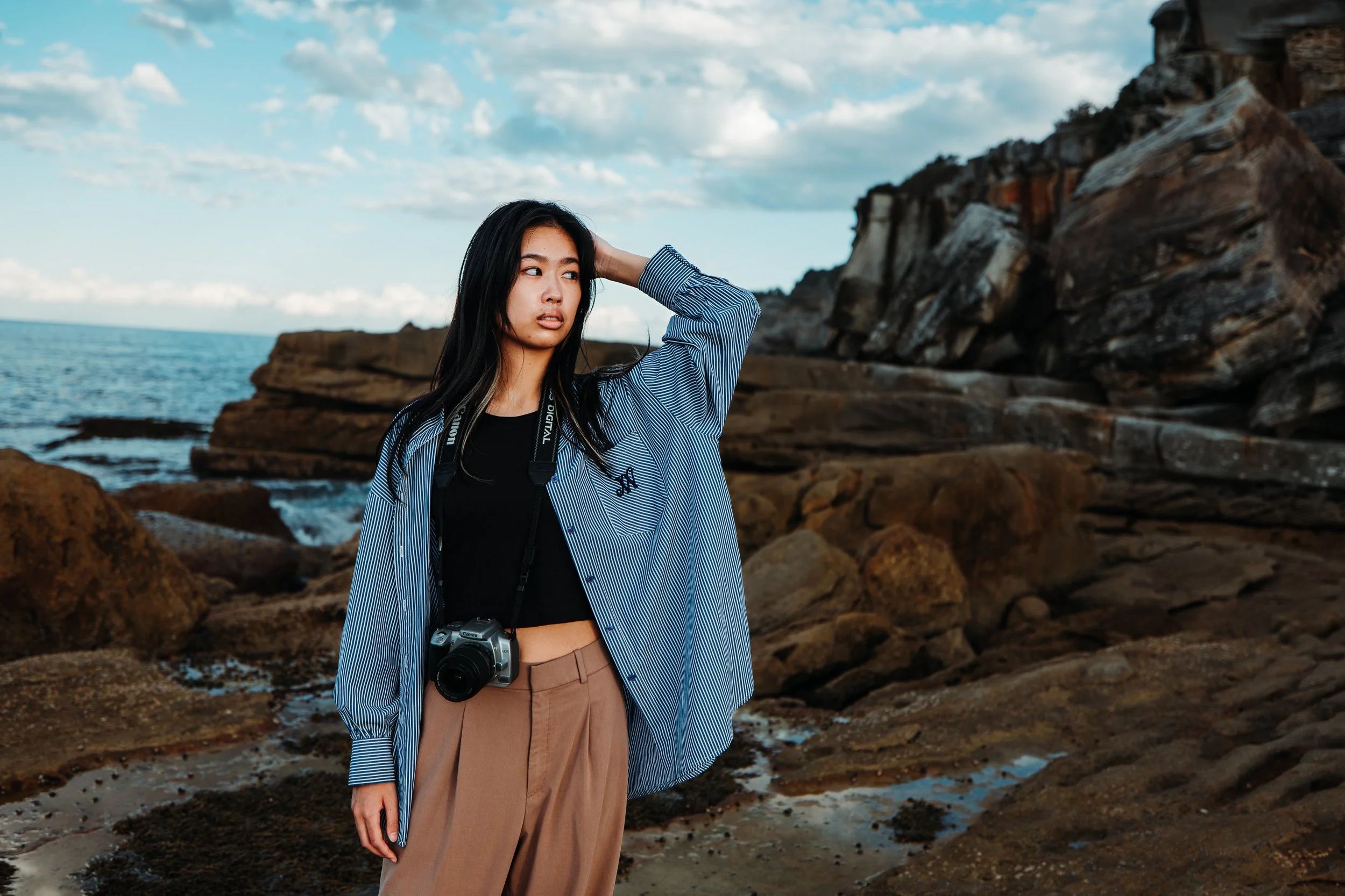 A young woman standing on a rocky coastline with the ocean and cloudy sky in the background. She is wearing a loose striped shirt over a black top, beige pants, and has a camera hanging around her neck, with her hand resting on her head, looking to t
