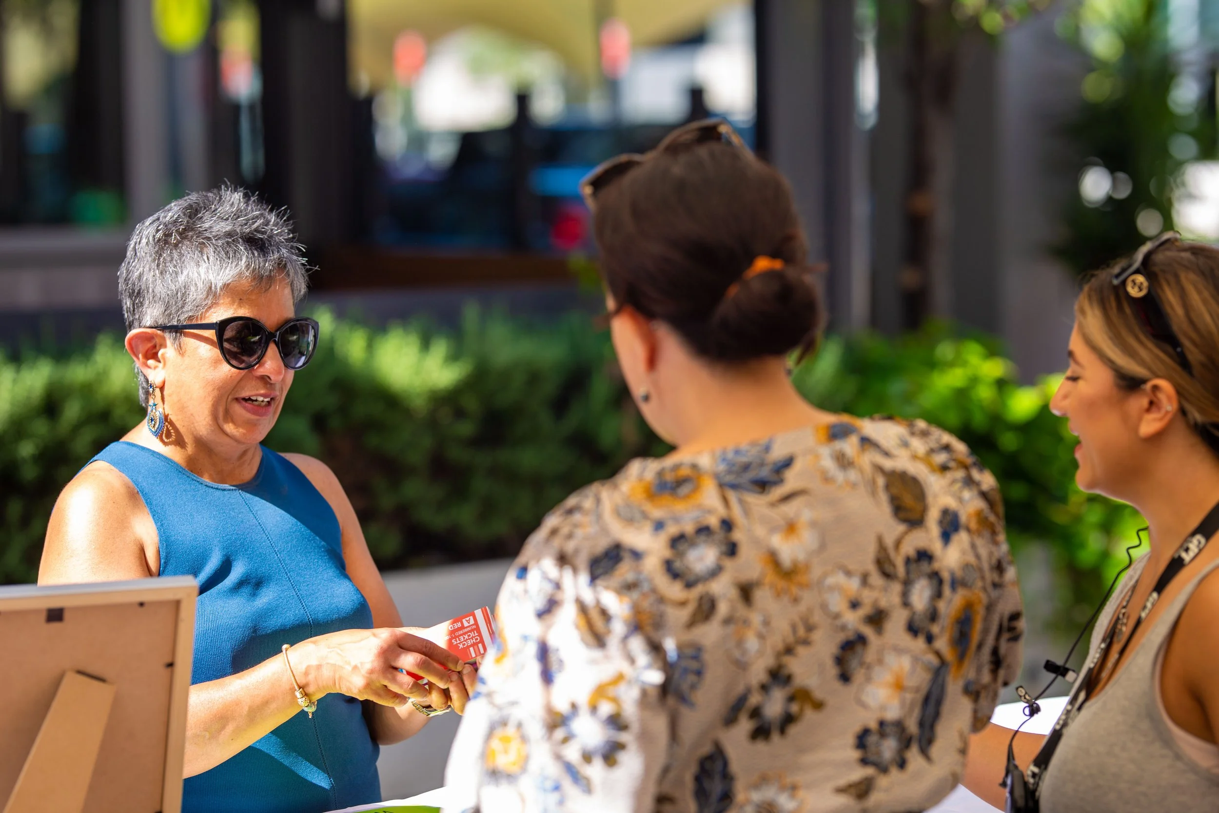 Three women engaged in conversation outdoors, with one woman holding a card, possibly at a restaurant or cafe.