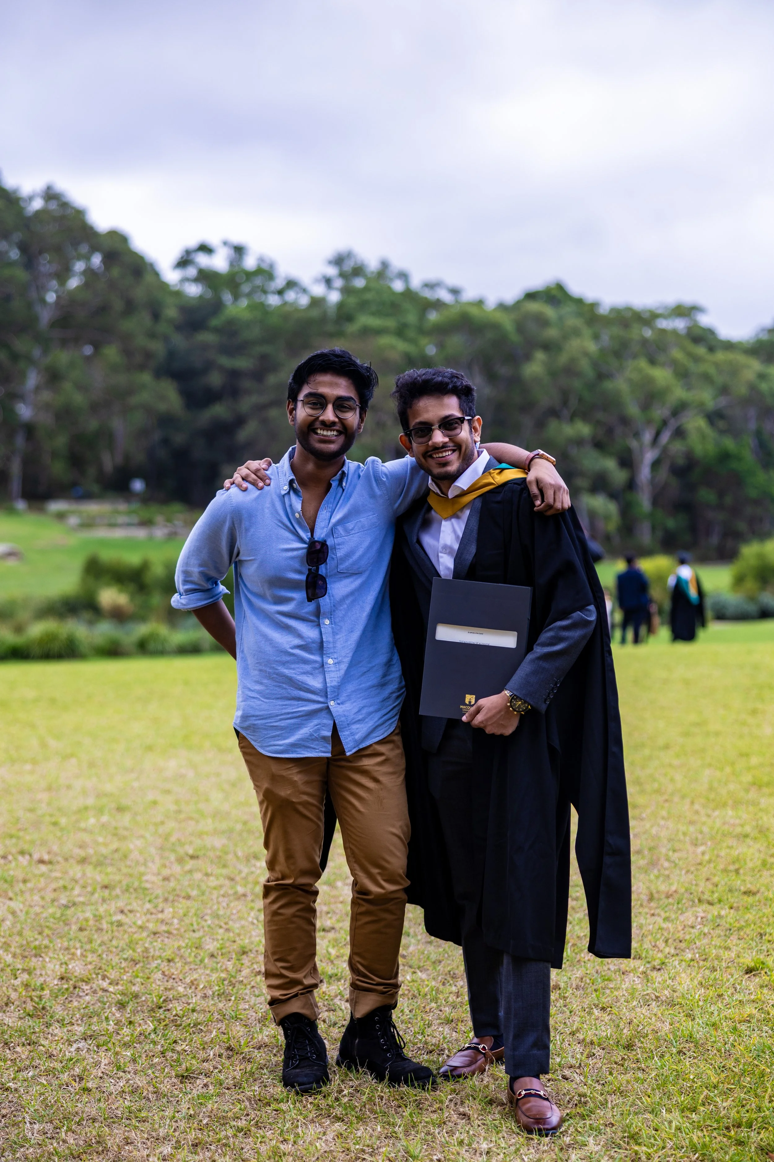 Two young men, one in casual attire and the other in a graduation gown and cap, smiling and posing together on a grassy area outdoors, likely celebrating a graduation.