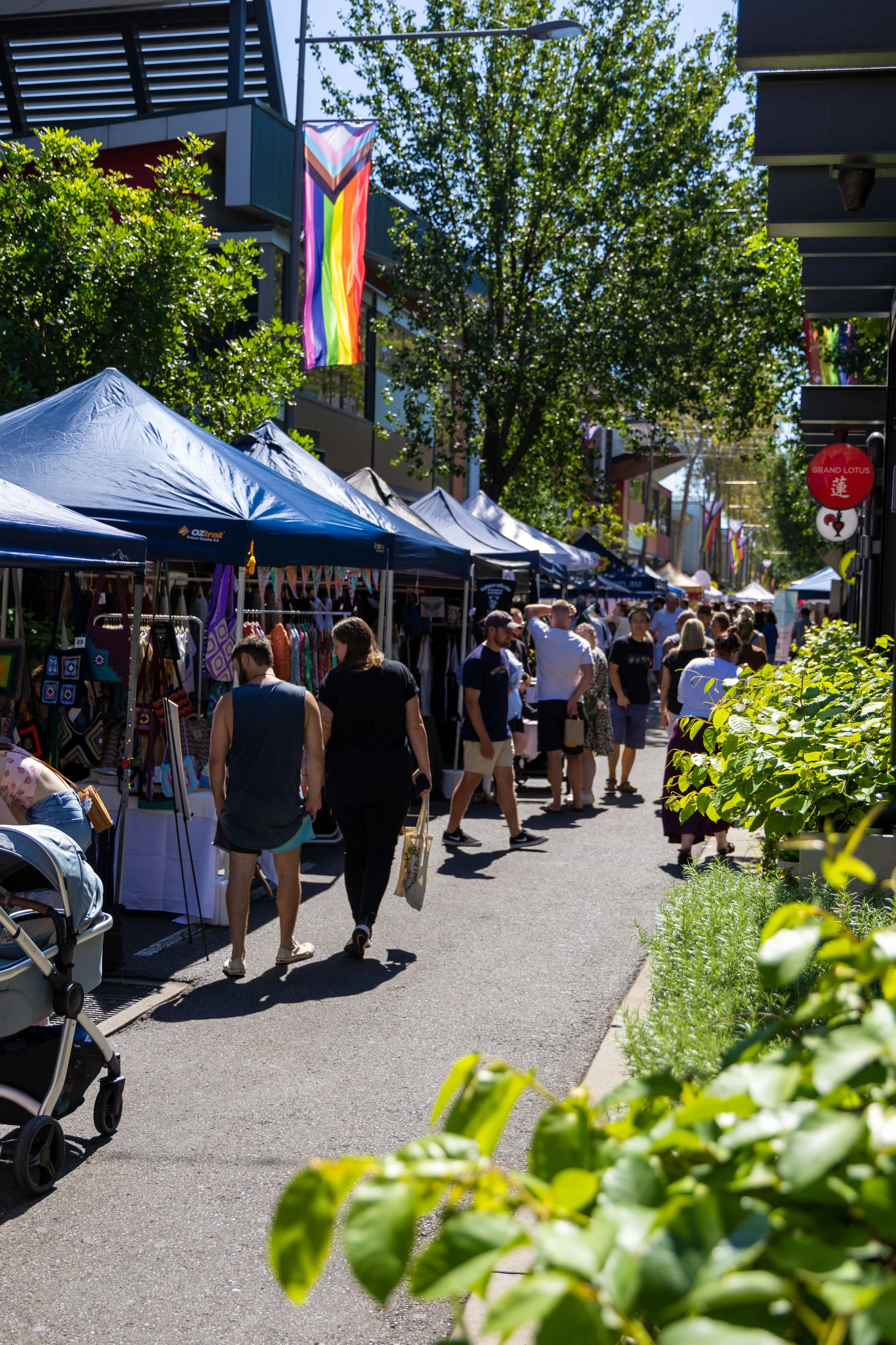 People strolling along a lively outdoor market with vendor booths and colorful merchandise, decorated with rainbow pride flags under a bright sunny sky.