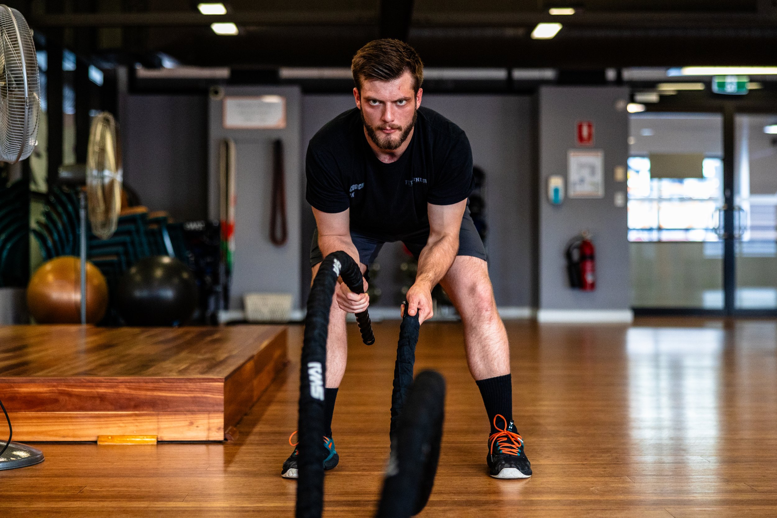 A young man in black athletic clothes engages in battle rope exercises in a gym.