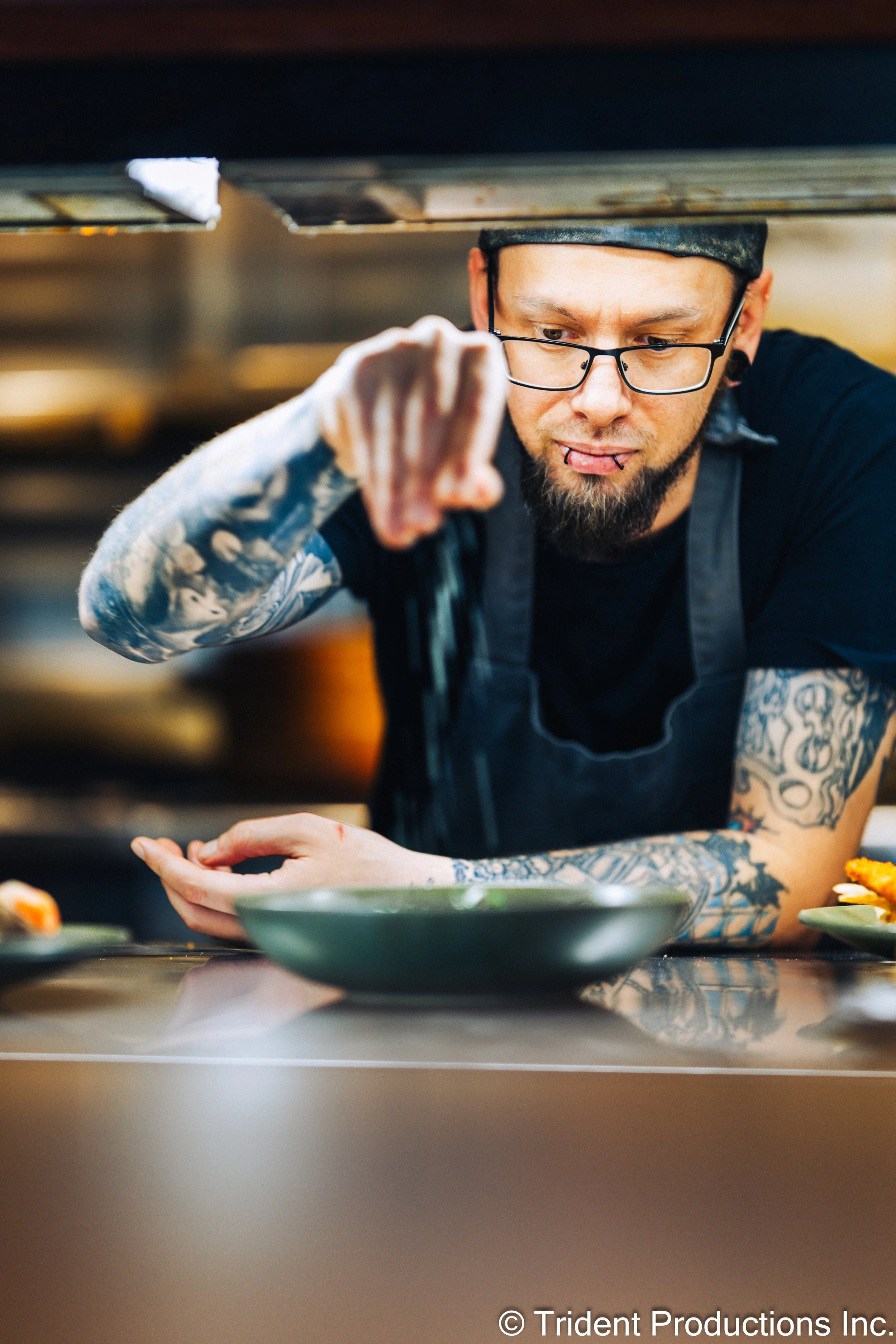 A tattooed chef with glasses and a beard, wearing a black apron, is seasoning a dish in a commercial kitchen.