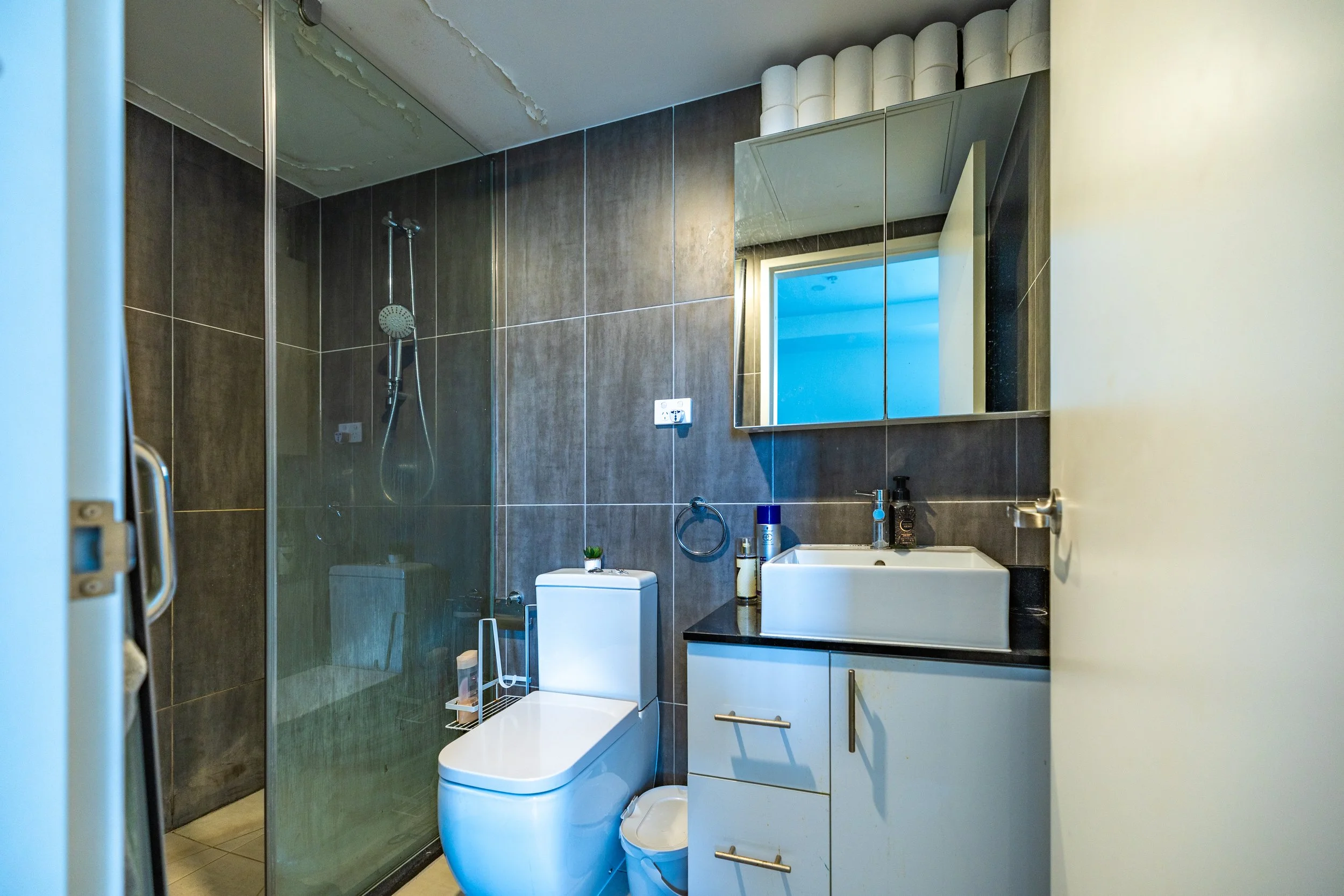 Small modern bathroom with a walk-in shower, white toilet, black countertop sink, and a wall-mounted mirror. Bathroom items on the counter include hand soap, lotion, and a can of spray. The wall behind the sink features dark tiles, and a small ceilin