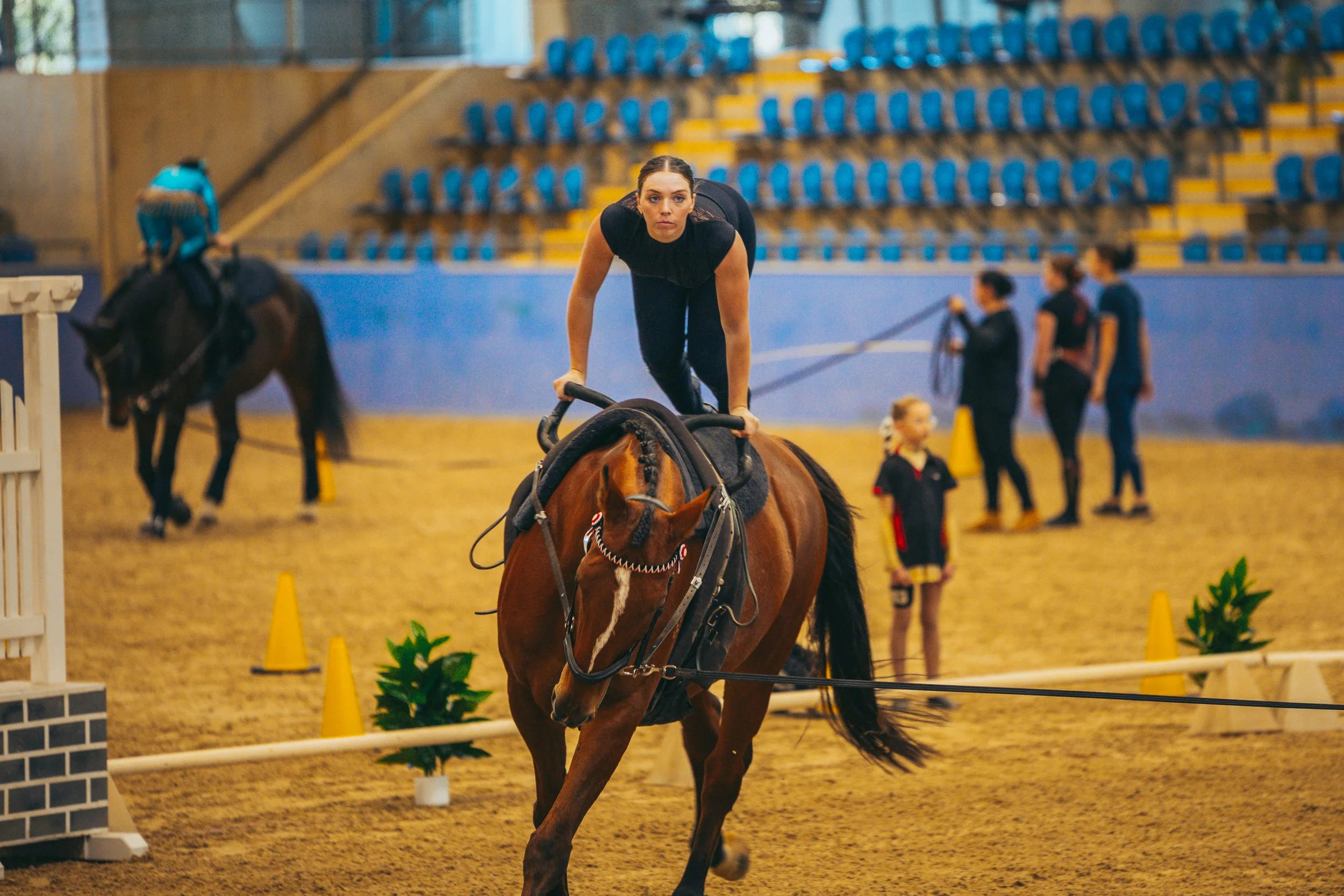 A woman riding a horse inside an indoor riding arena, with other people and horses in the background and safety cones on the ground.