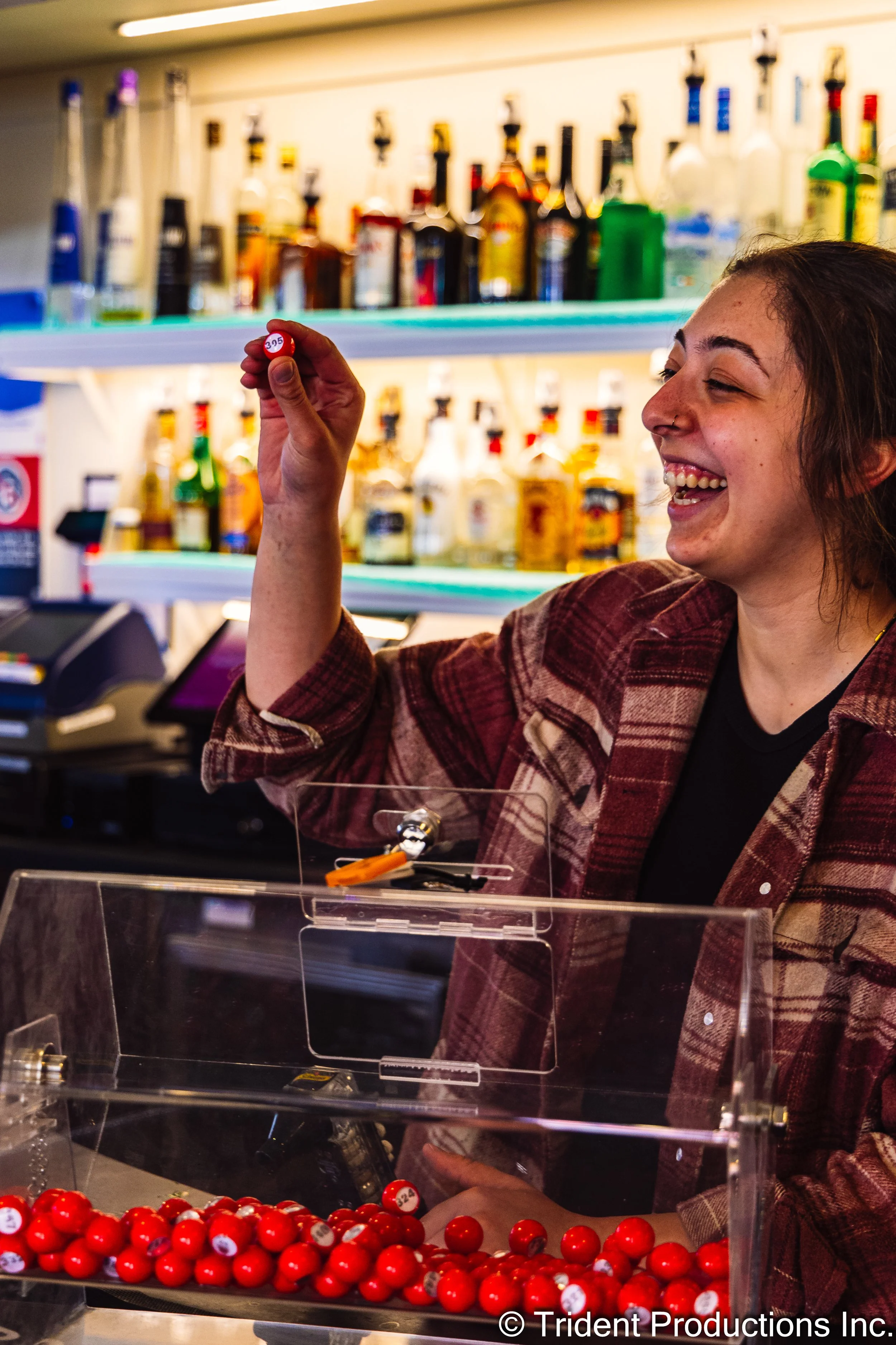 A smiling woman in a plaid shirt at a lottery ticket counter, holding a red lottery ball with the number 305.