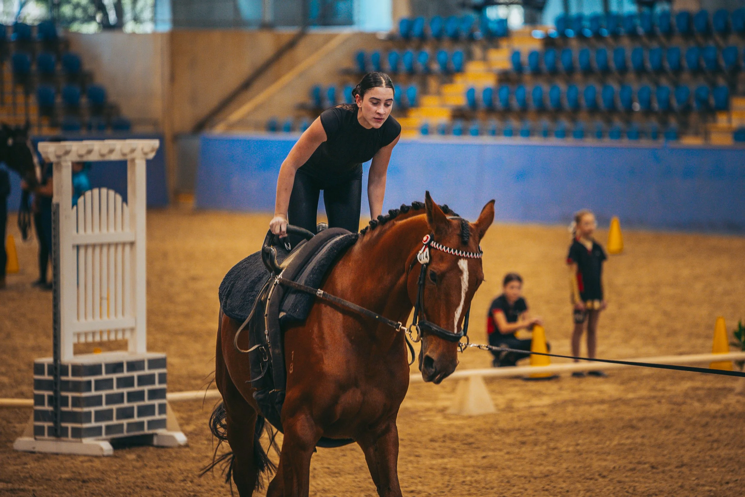 A young woman riding a horse in an indoor equestrian arena, with two young people sitting on the ground and yellow cones in the background.
