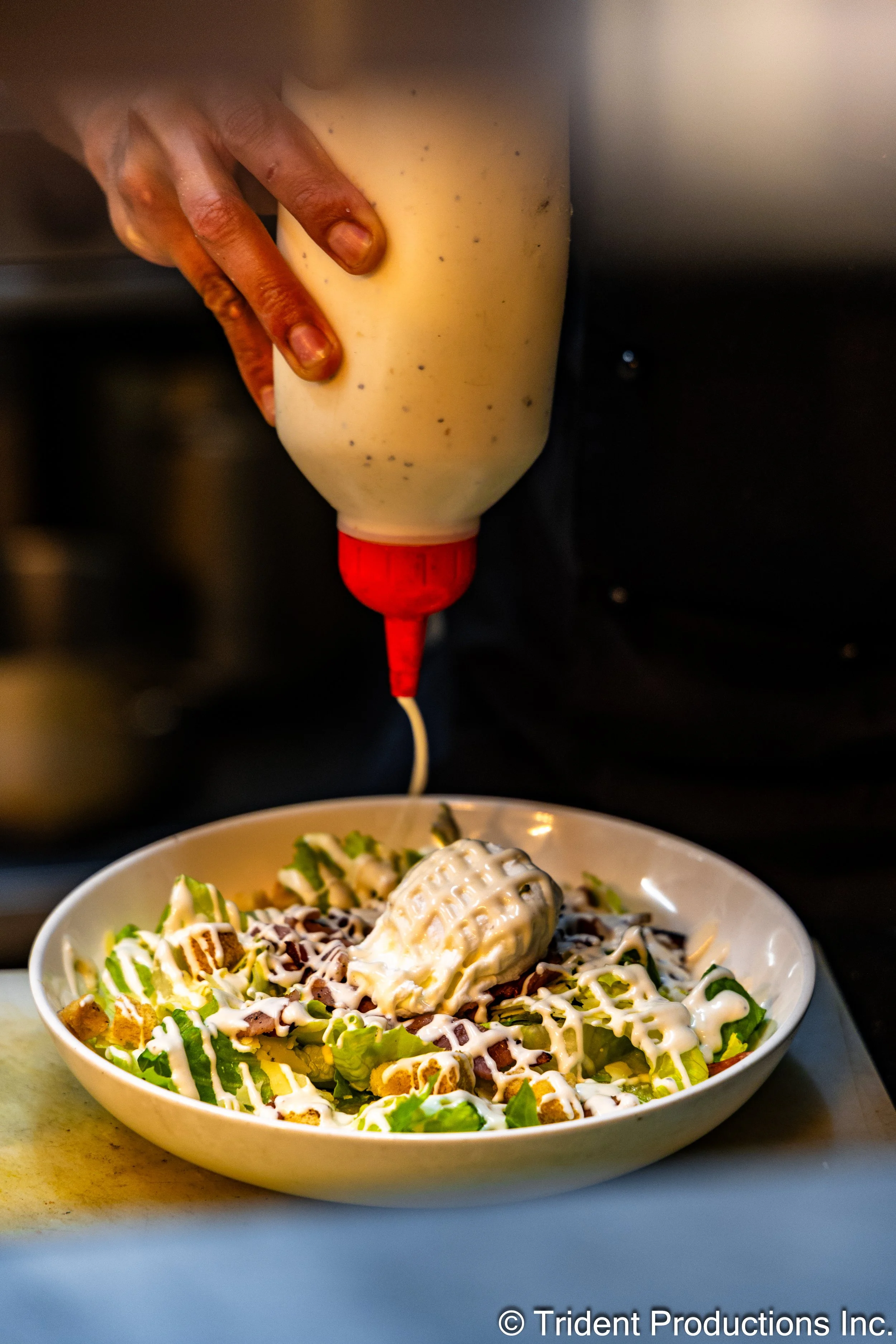 A person drizzling creamy salad dressing from a squeeze bottle onto a salad in a white bowl, containing lettuce, diced vegetables, shredded cheese, and a dollop of dressing.