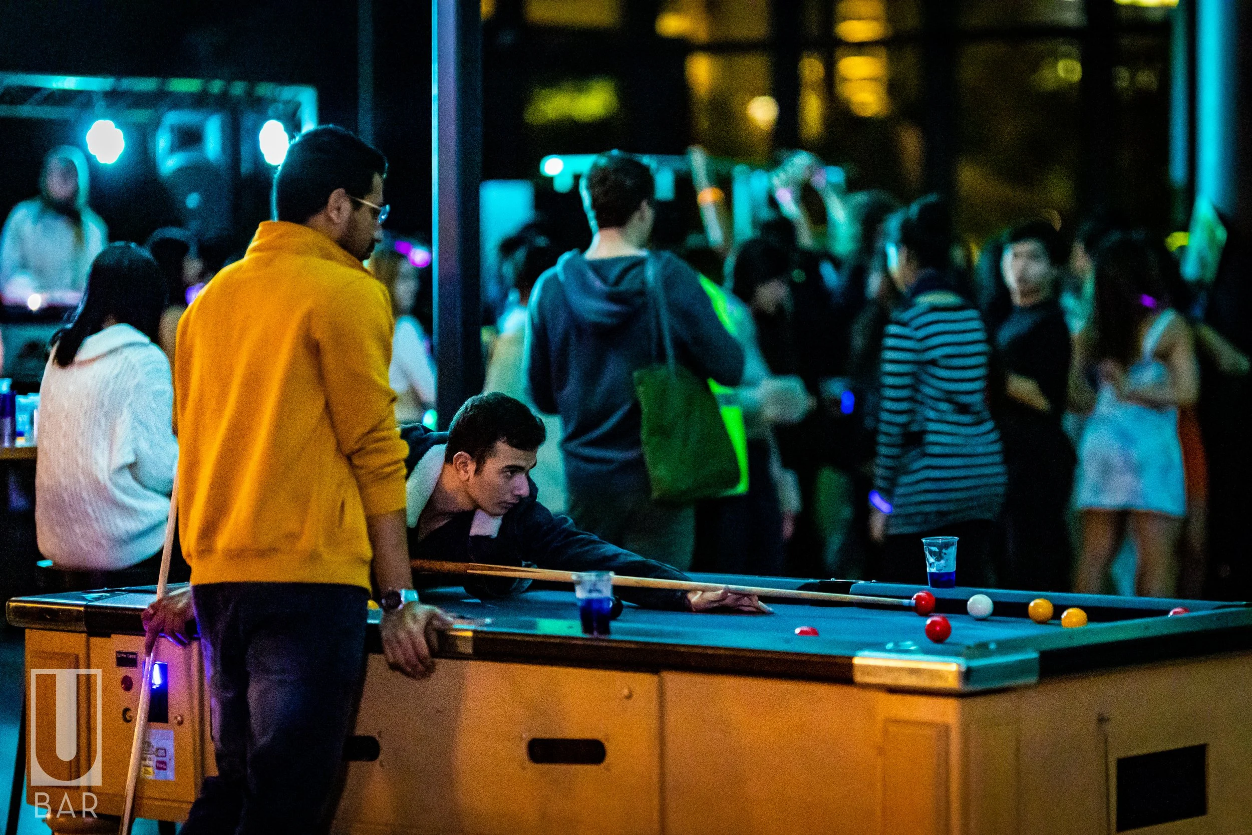 Two young men playing pool at a bar, with a crowd of people in the background, illuminated by blue and green lighting.