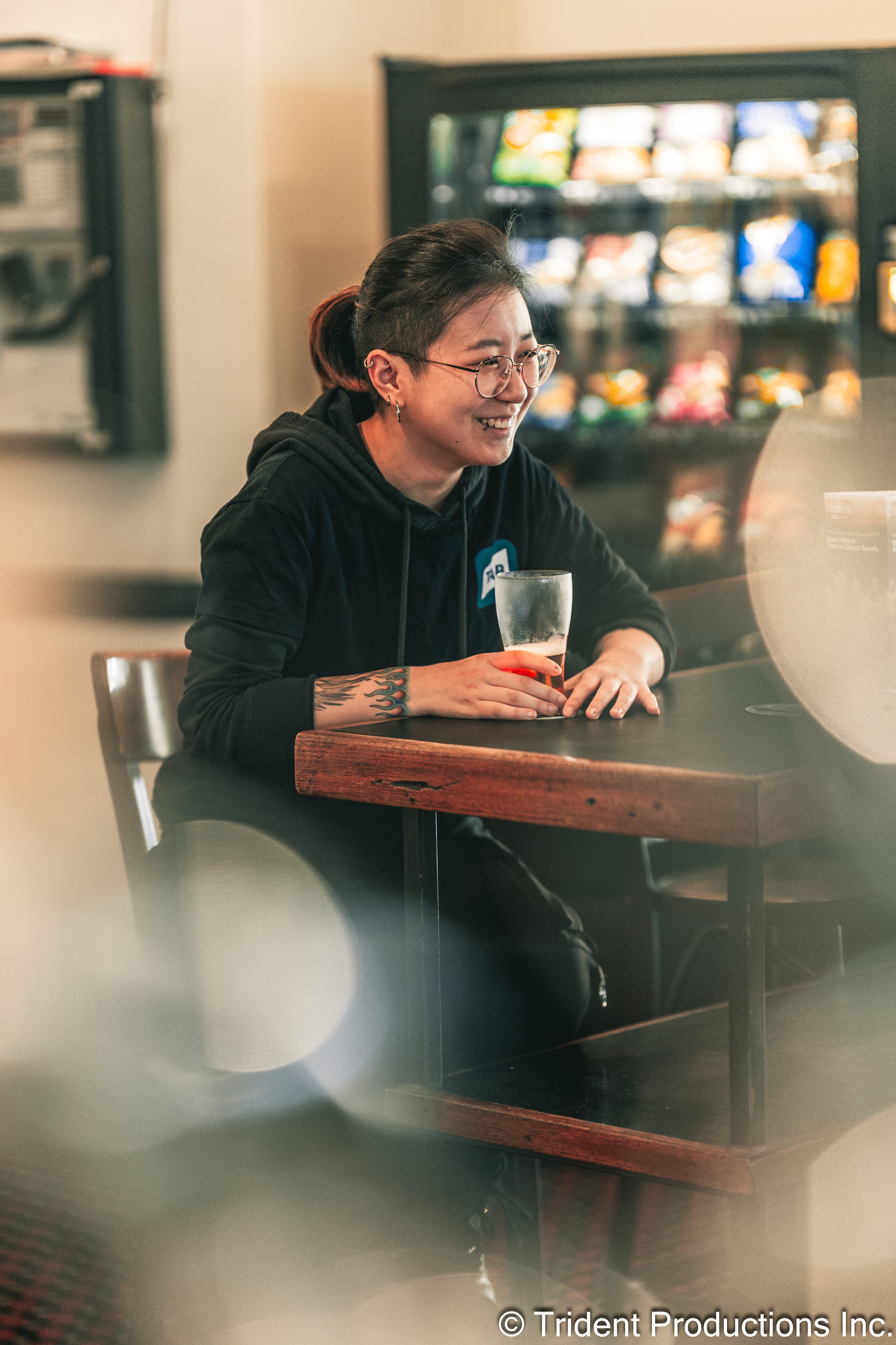 A woman with glasses and tattoos on her arm sitting at a table in a casual setting, holding a glass of beer and smiling.
