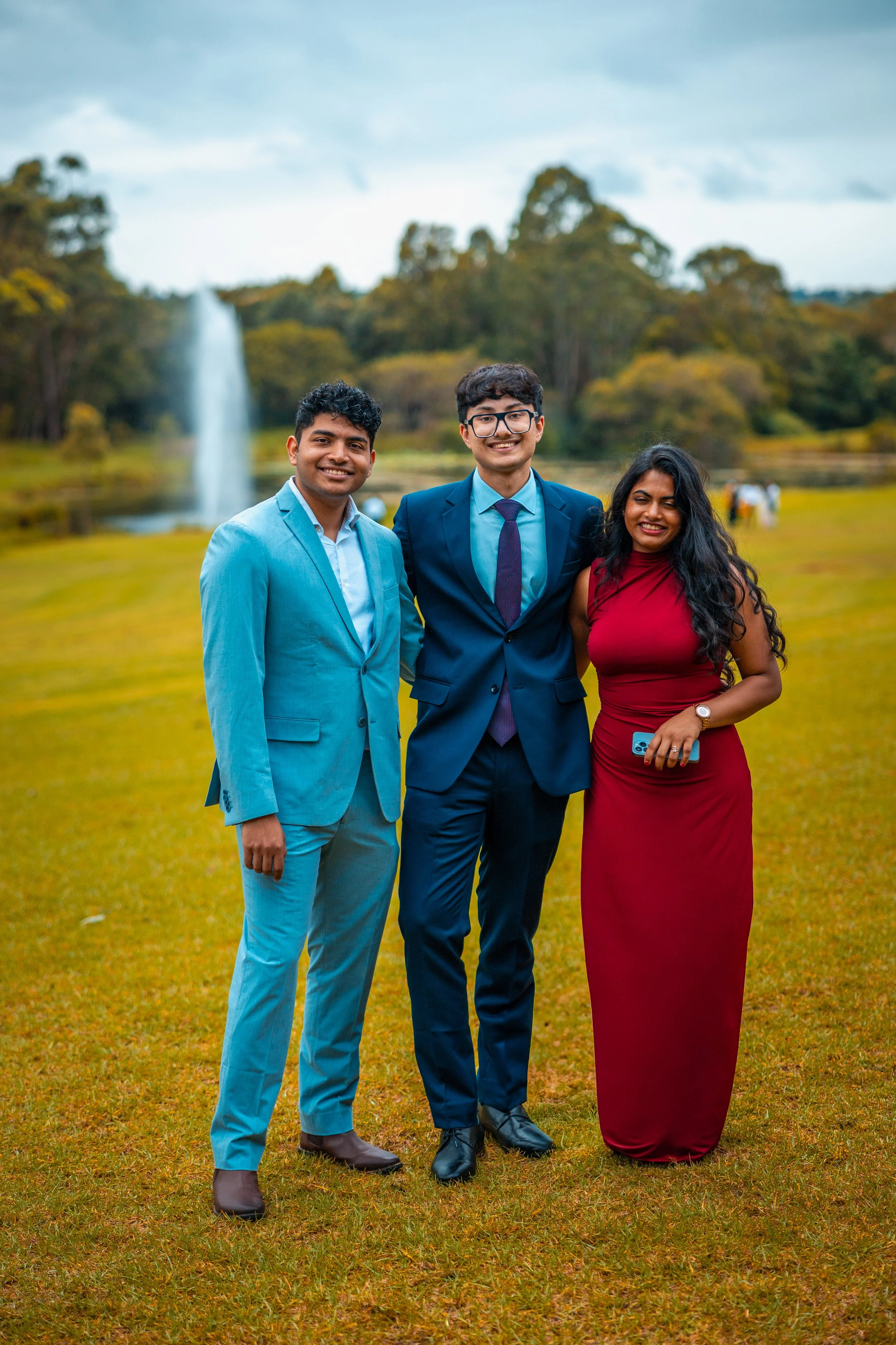 Three people dressed in formal attire standing outdoors on grass with a fountain and trees in the background, smiling at the camera.