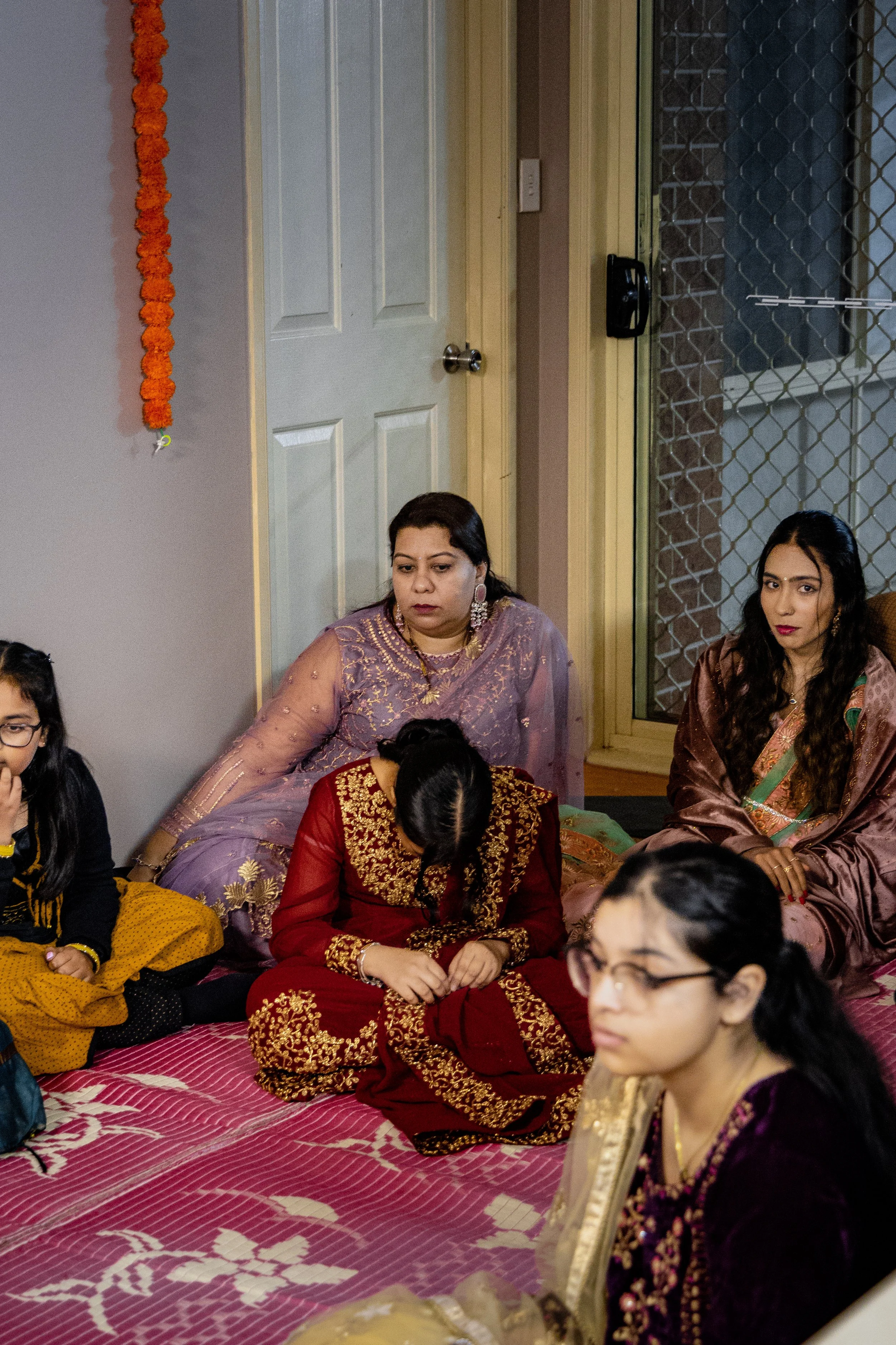 Group of women and a girl sitting on a pink patterned mat in a room, dressed in Indian traditional clothing, with a door and window in the background.