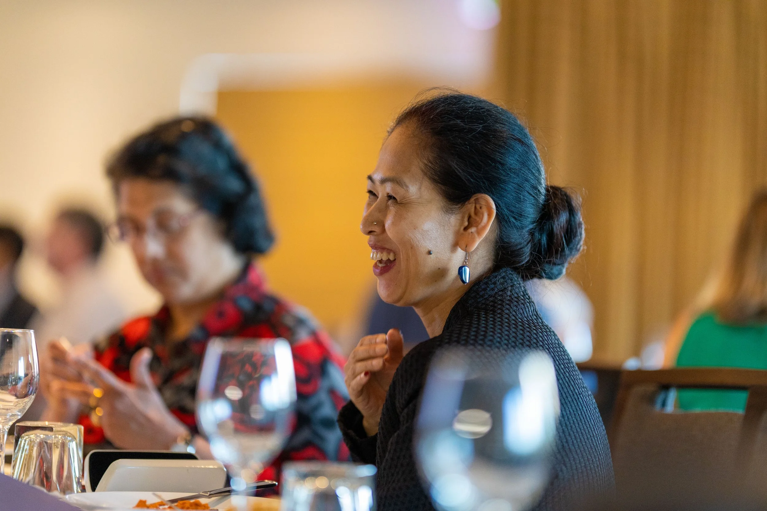 Smiling woman with black hair styled in a bun, wearing blue earrings and a black textured blazer, sitting at a dining table with wine glasses and plates, engaged in conversation at a social event.