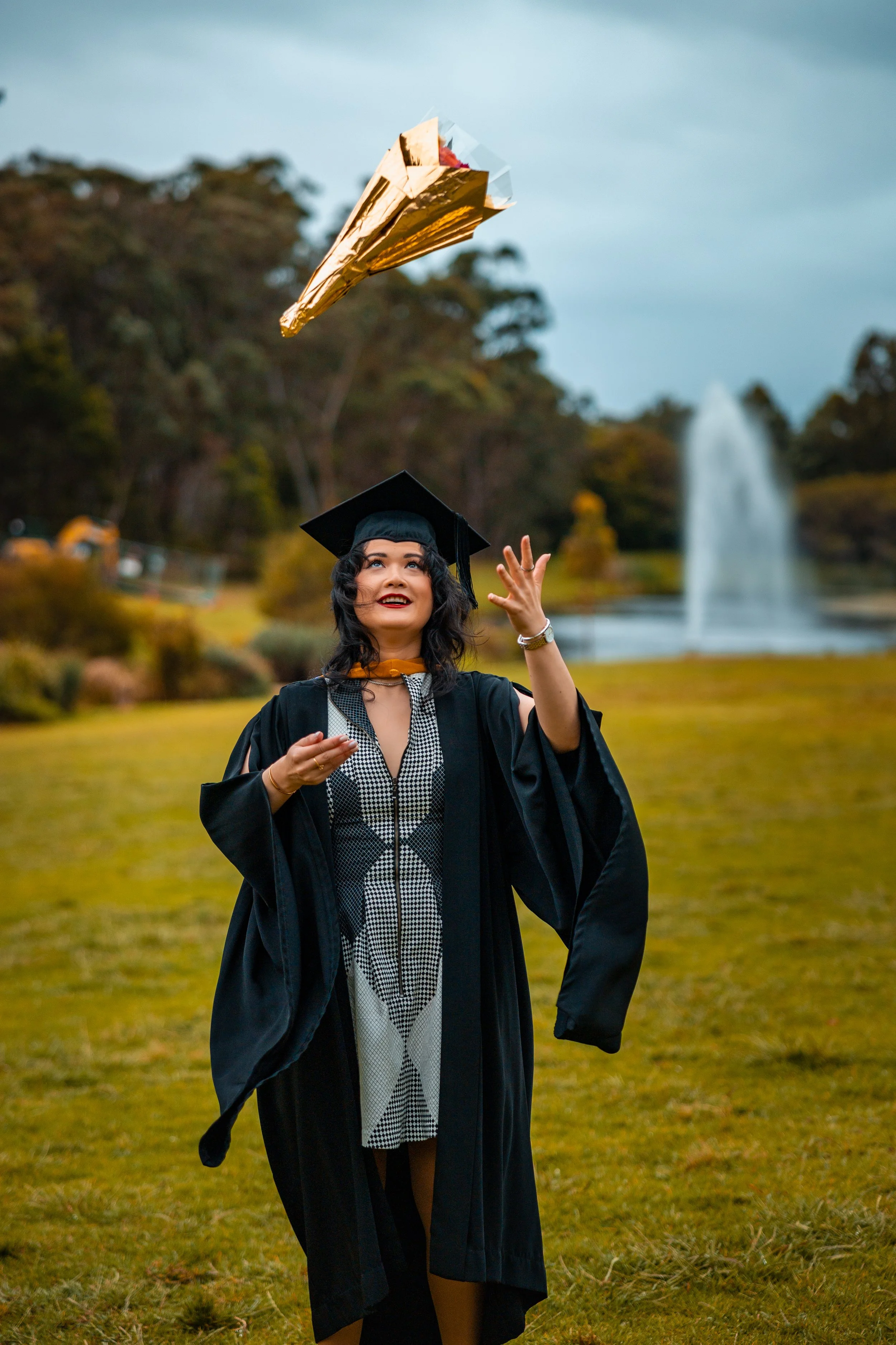 A young woman in a graduation cap and gown is outdoors on a cloudy day, tossing her diploma tube into the air and smiling.