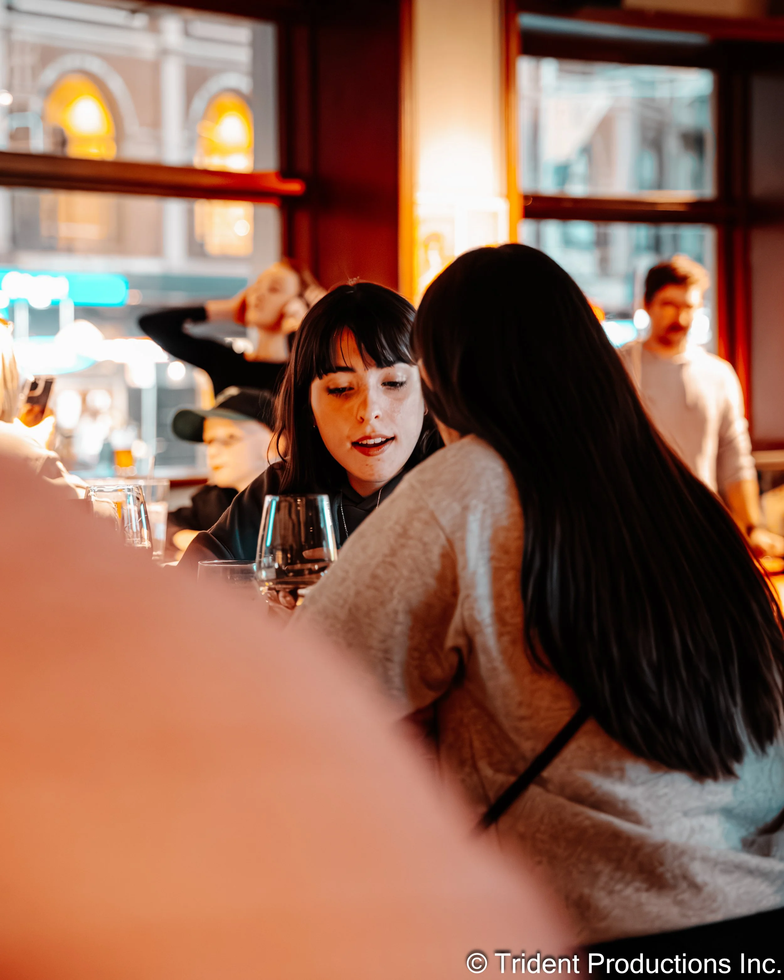 Two young women are seated at a table in a warmly lit restaurant or cafe, engaged in conversation. One has black hair with bangs, and the other has long dark hair. In the background, other patrons are visible, including a woman with blonde hair and a man with glasses, with large windows allowing natural light to fill the space.
