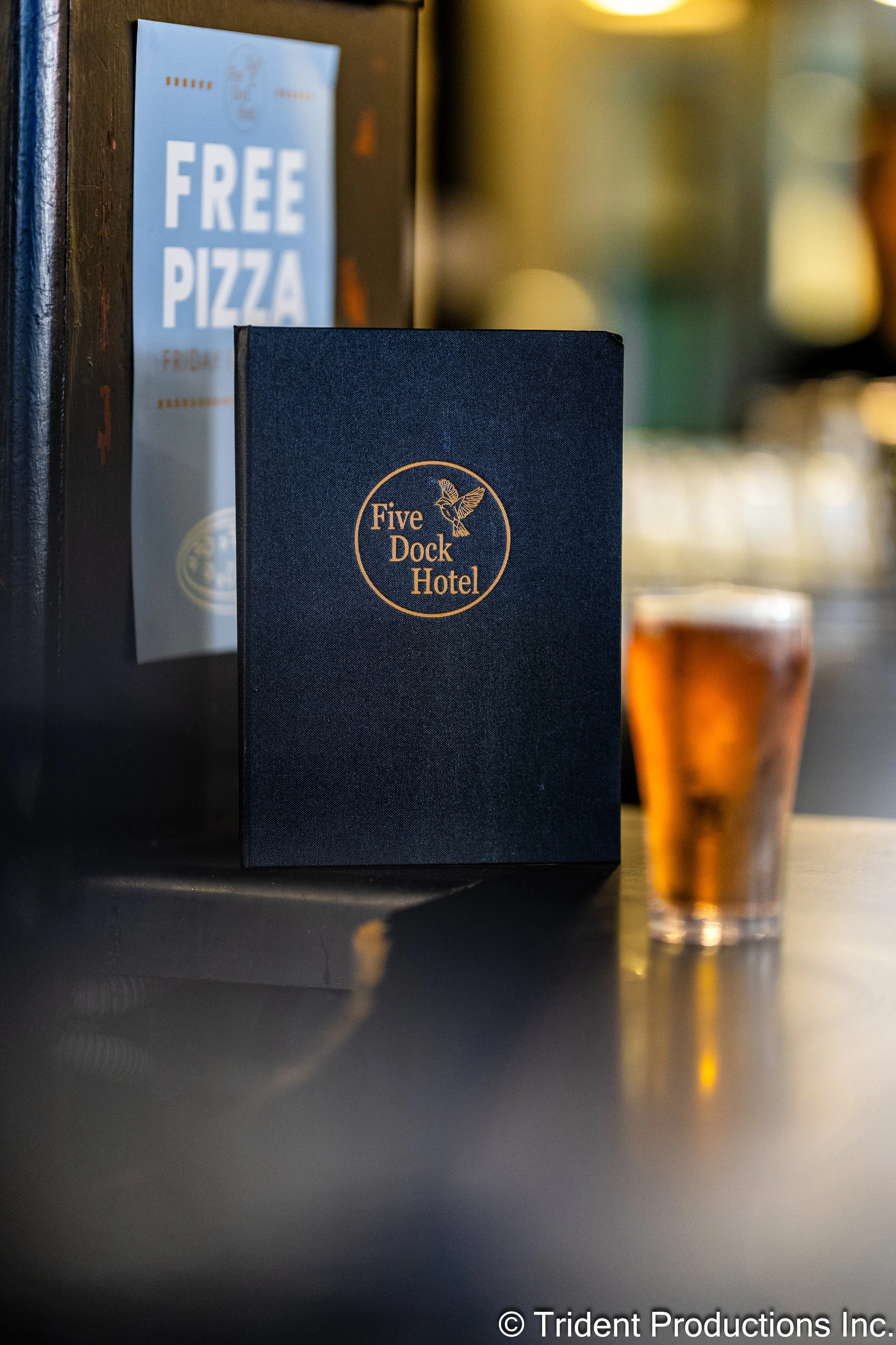 Menu or brochure from Five Dock Hotel, a dark-colored menu with gold lettering and a logo featuring a bird, on a bar counter, with a glass of beer or cider with a golden amber color nearby.