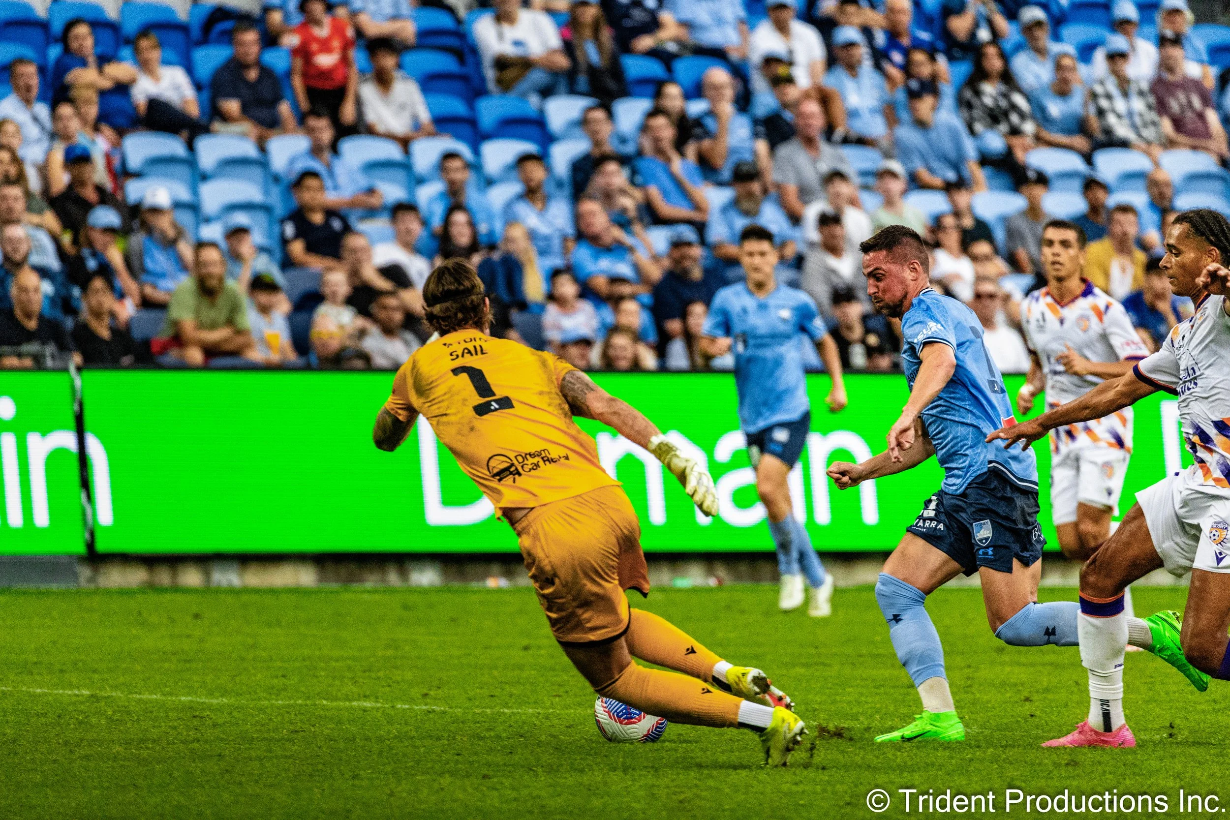 A soccer match in progress with players trying to score. The goalkeeper in yellow is attempting to block the shot by kicking the ball. The players are wearing blue and white jerseys, and spectators are watching from the stands.