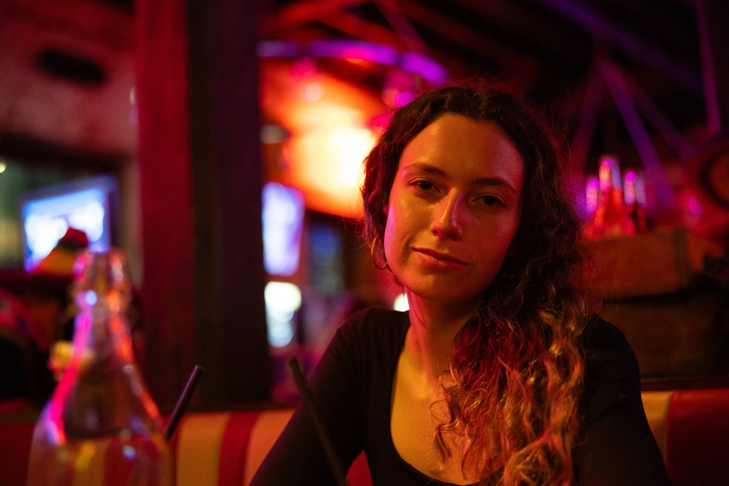 A young woman with long curly hair sitting in a dimly lit restaurant or bar with colorful lights in the background.