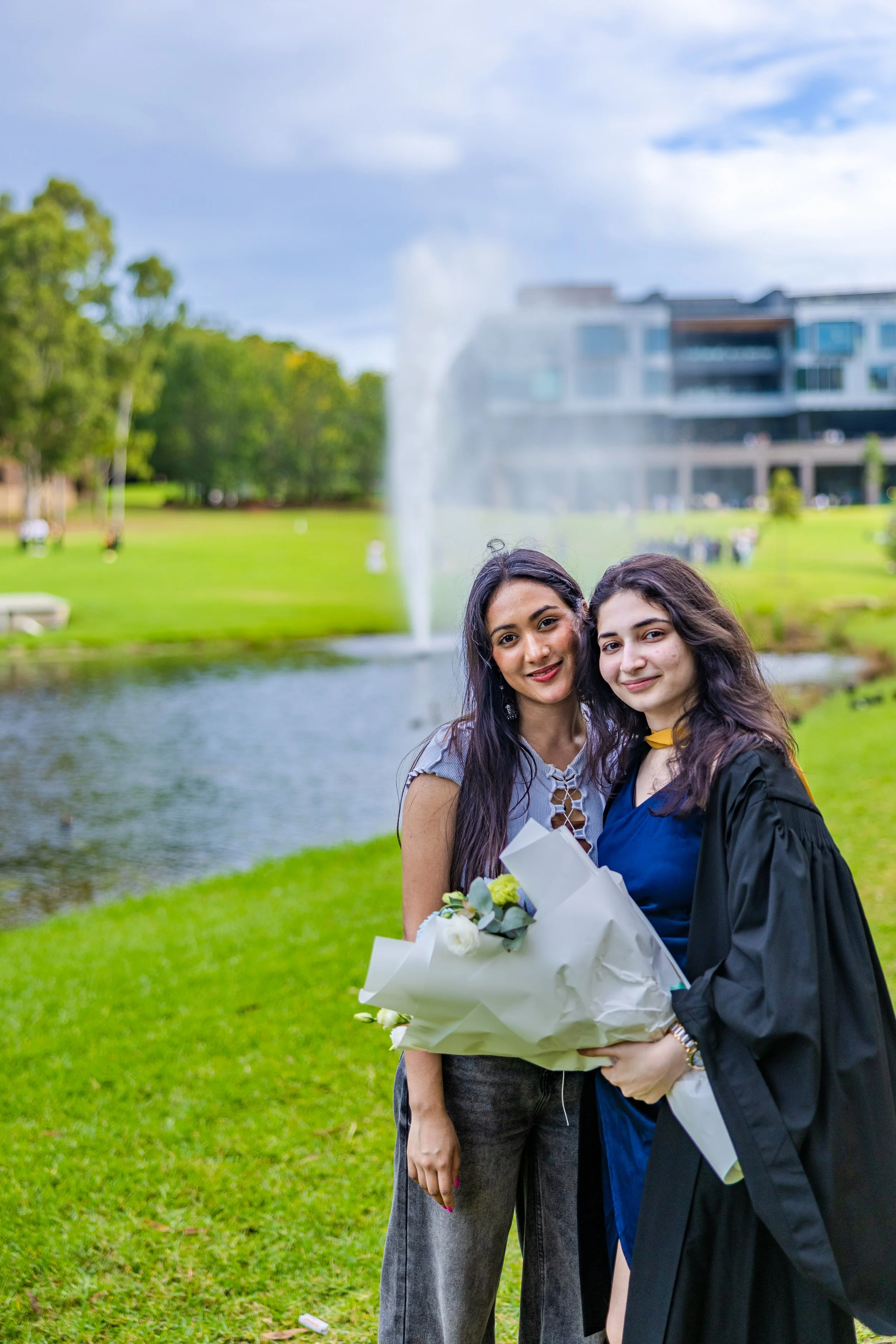 Two women, one in casual attire and the other in graduation gown, smiling and holding a bouquet of flowers, standing near a pond with fountain, trees, and modern building in the background.