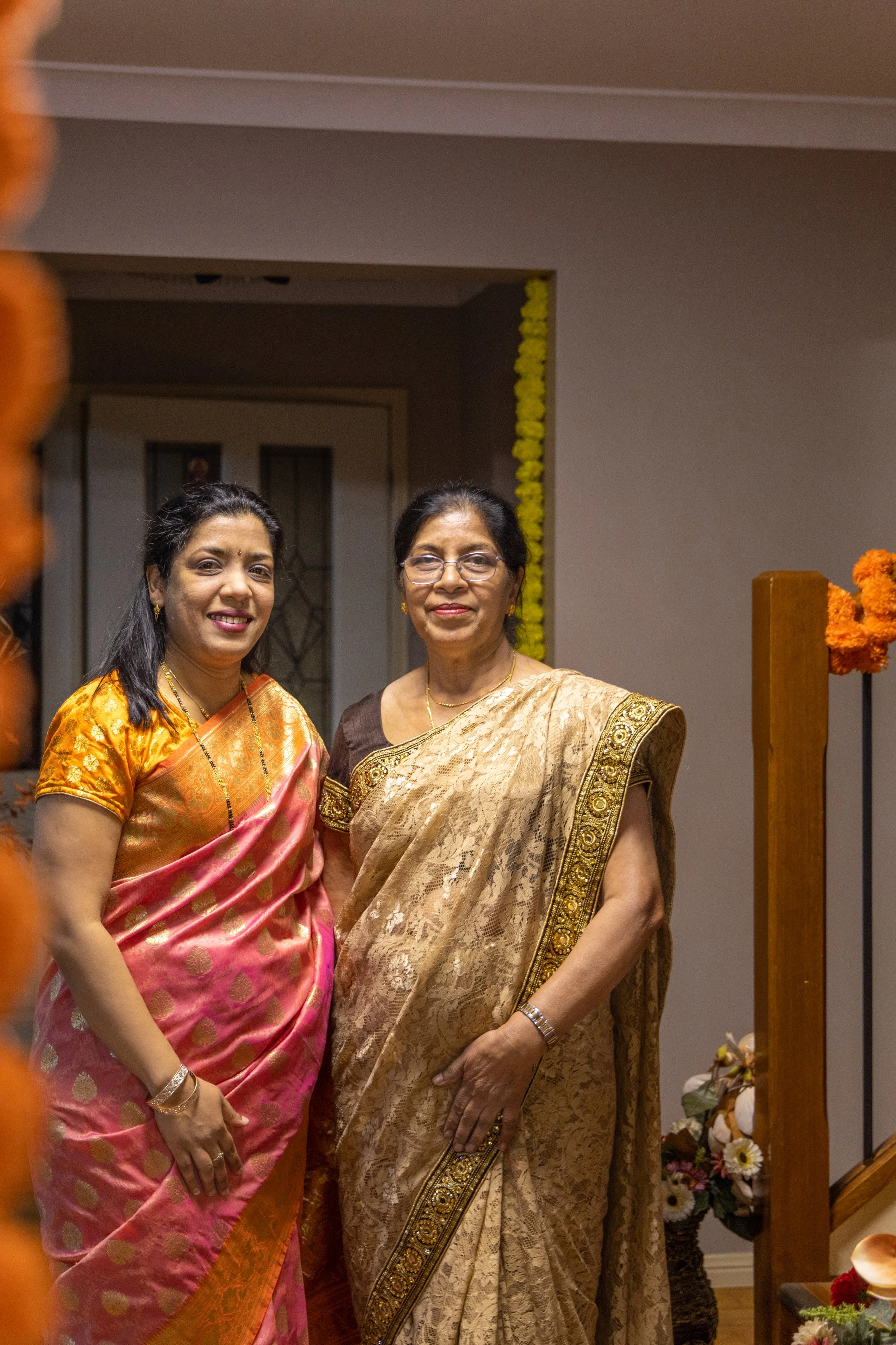 Two women in traditional Indian sarees standing together indoors, smiling at the camera, with floral decorations in the background.
