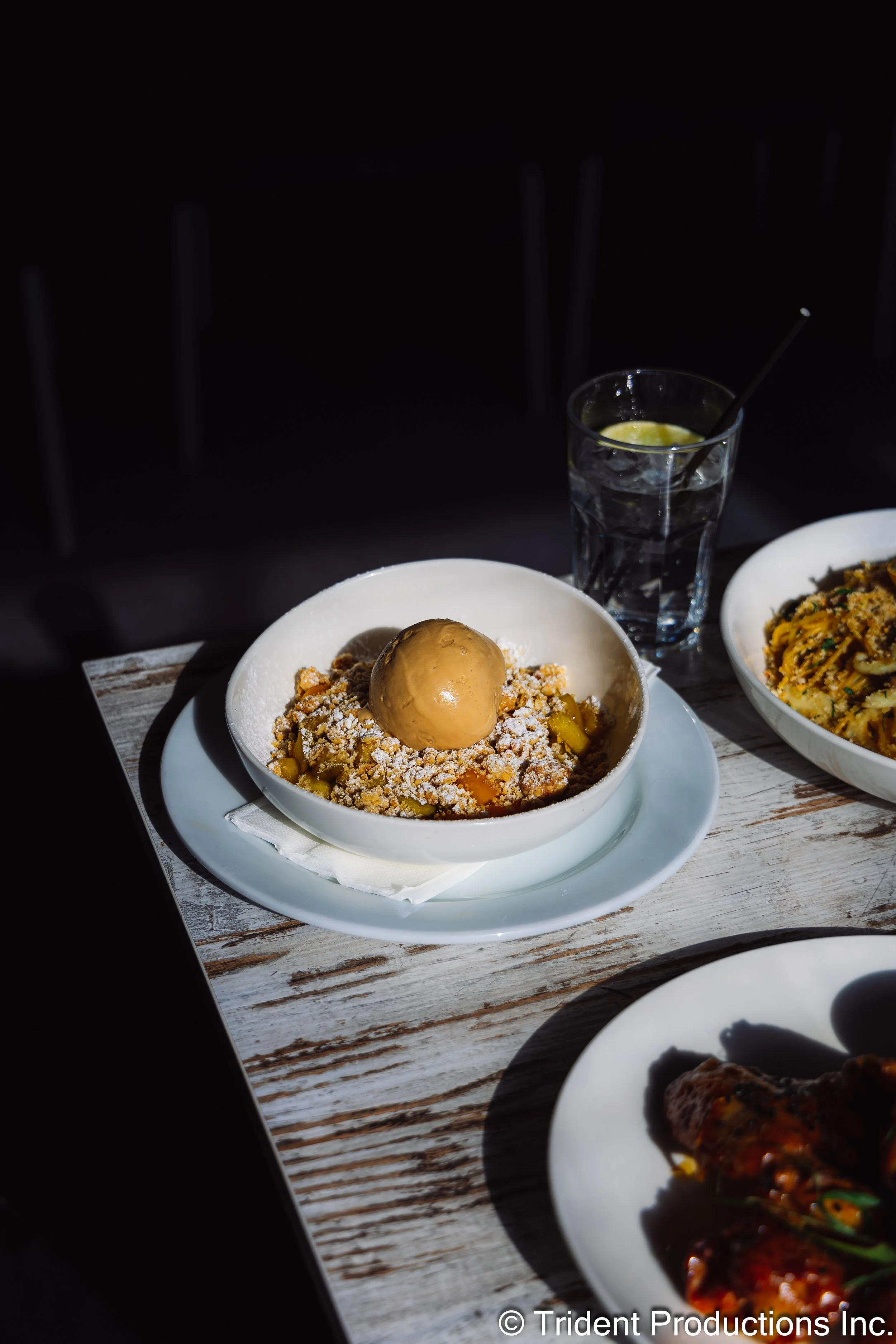 Dessert featuring a scoop of caramel ice cream with crushed cookies and yellow fruit pieces in a white bowl with a white plate underneath, on a wooden table.