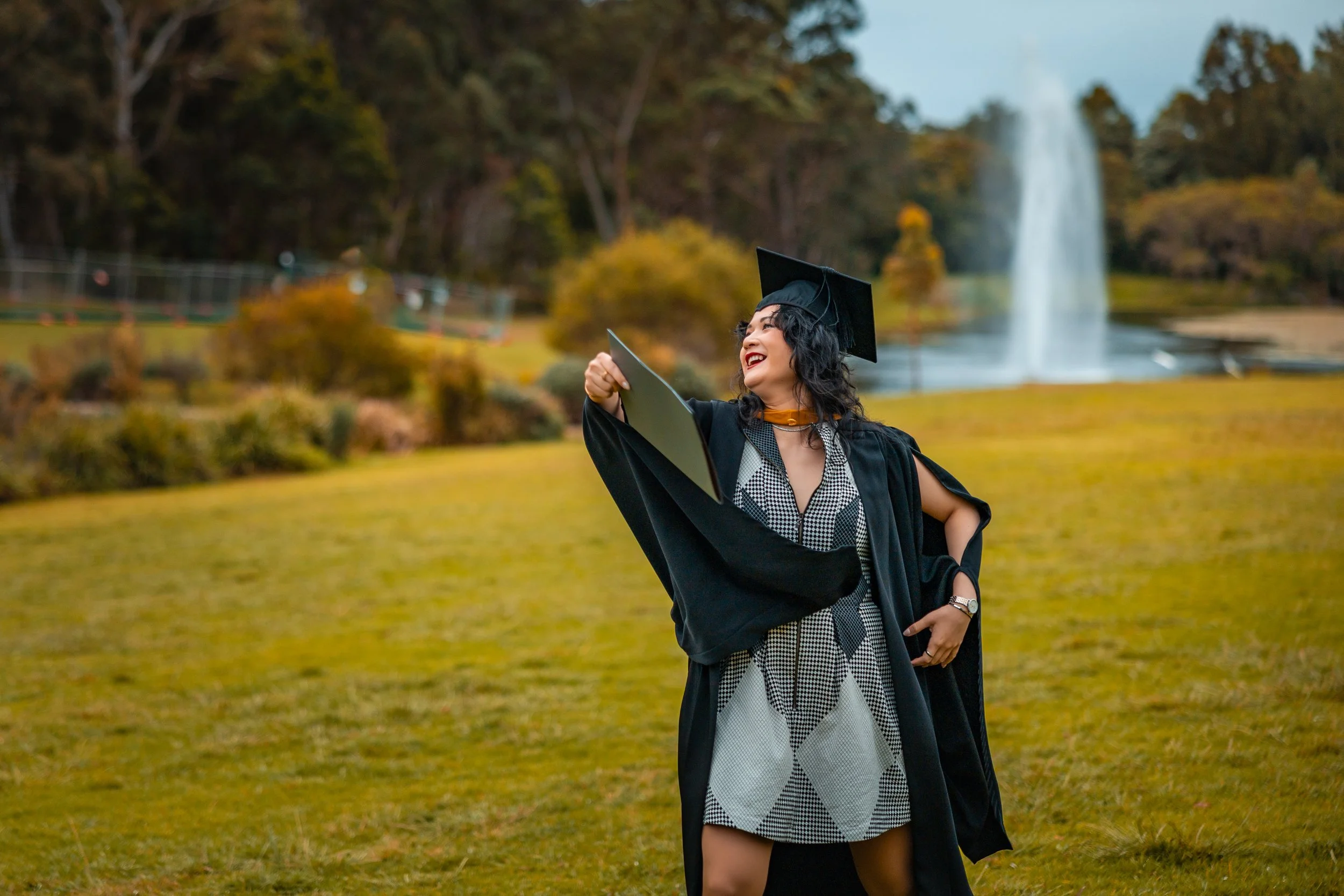 A woman in a graduation gown and cap holding her diploma, standing in a park with a fountain in the background.