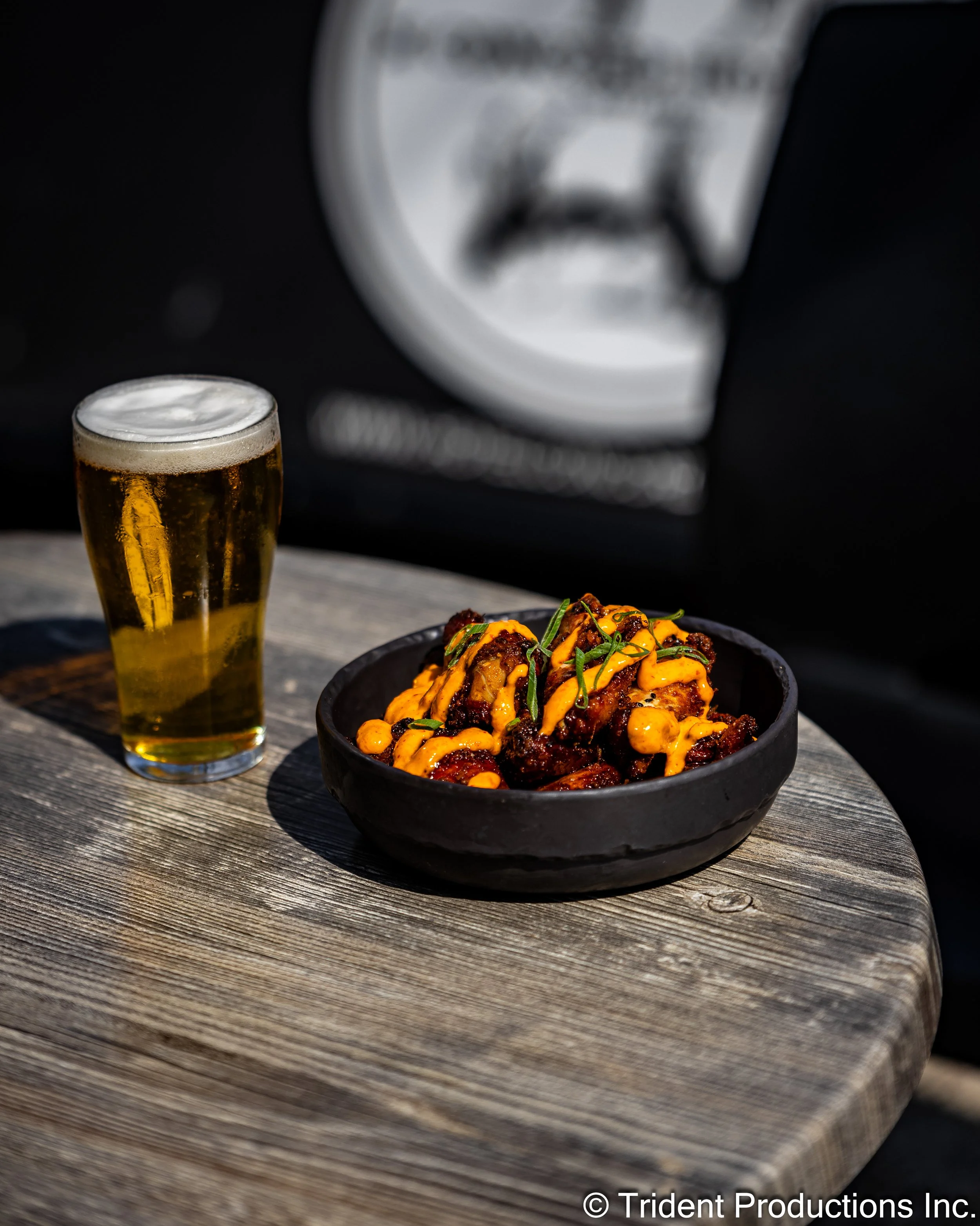 A black bowl filled with spicy chicken wings drizzled with orange sauce, topped with chopped green herbs, on a weathered wooden table. Next to the bowl is a pint glass of golden beer with a frothy head. The background is blurred and appears to be out