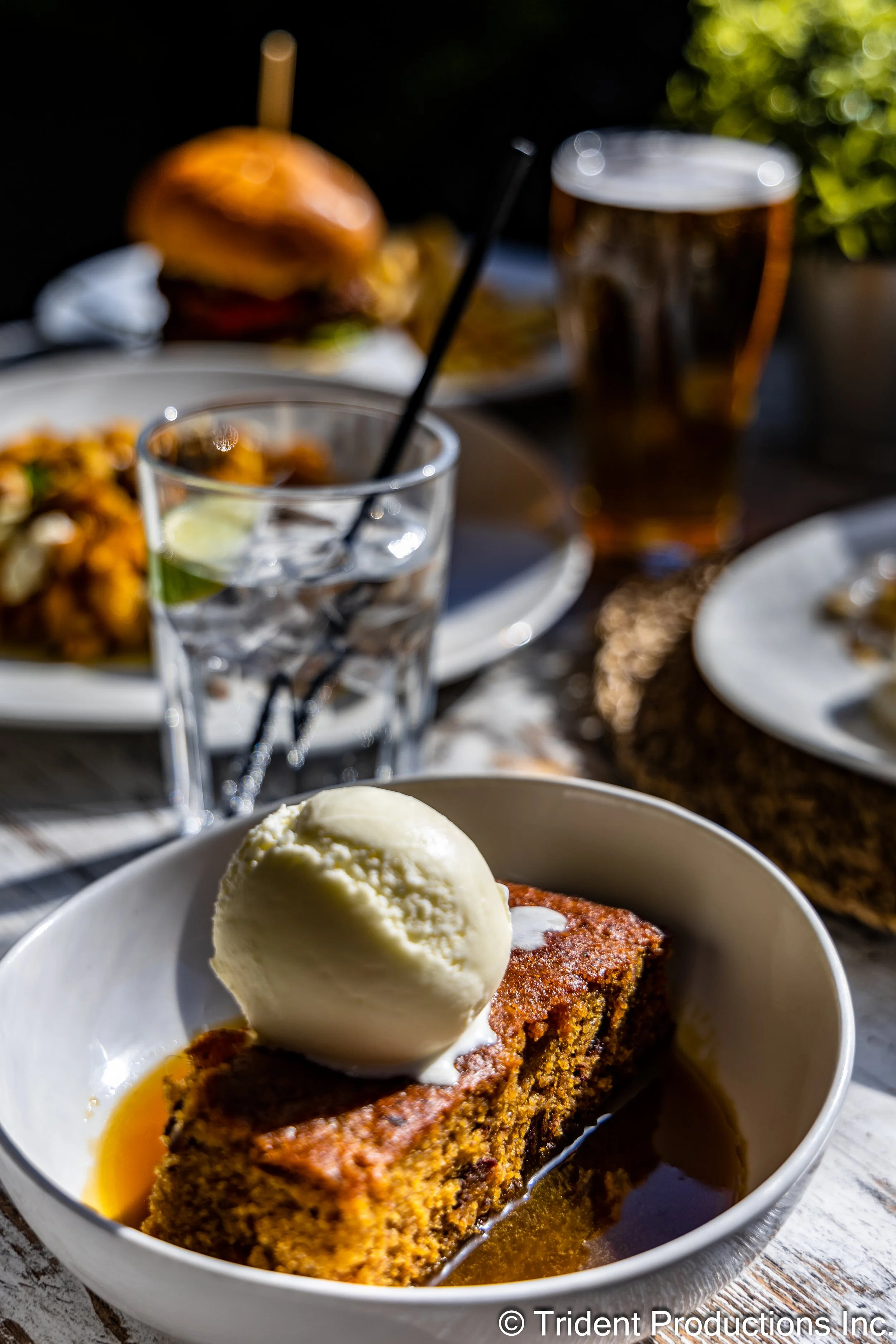 A slice of carrot cake with vanilla ice cream on top, served with caramel sauce in a white bowl, on an outdoor table with other plates of food, a glass of water with a lemon wedge, and a glass of beer in the background.