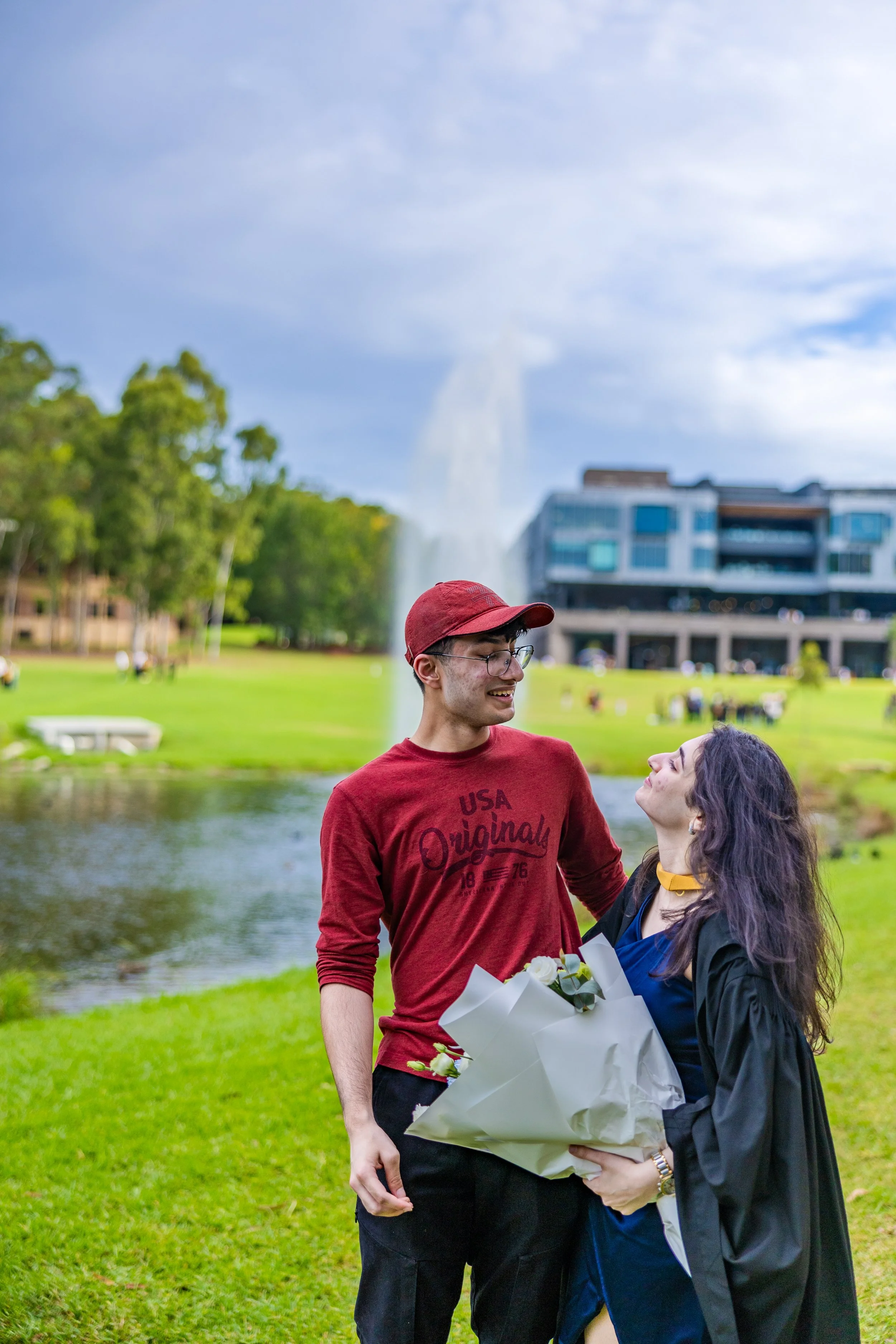 A young man and woman celebrating graduation at a park, with the woman holding a bouquet of flowers. The background features a pond, fountain, and modern buildings, under a partly cloudy sky.