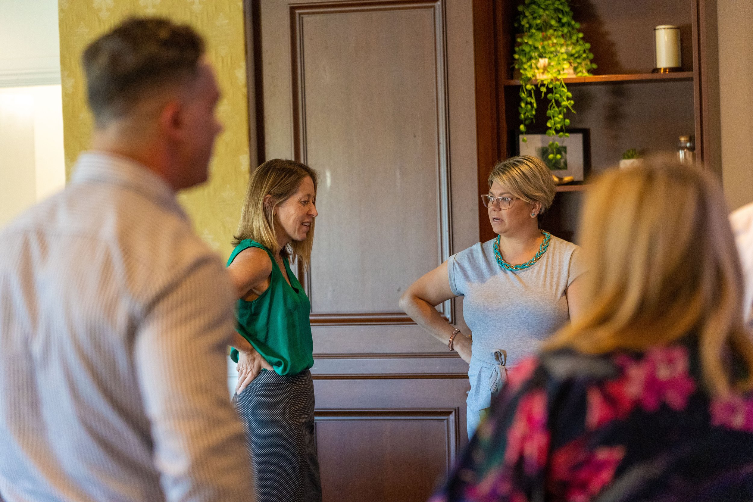 Two women and three blurred individuals engaged in conversation in a room, with shelves and potted plants in the background