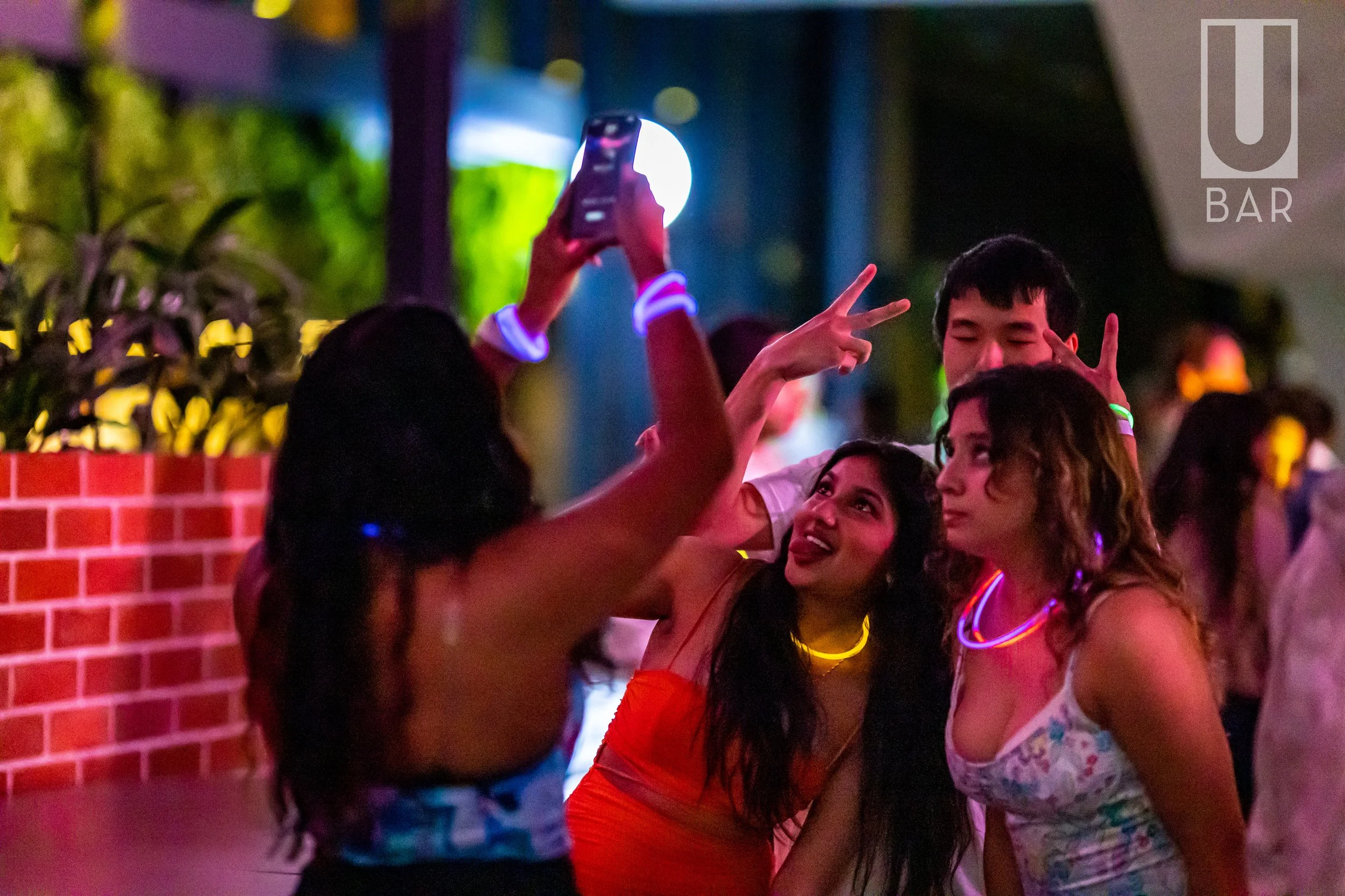 Four young people taking a selfie at a lively party, wearing glow necklaces and wristbands, with bright colorful lighting.