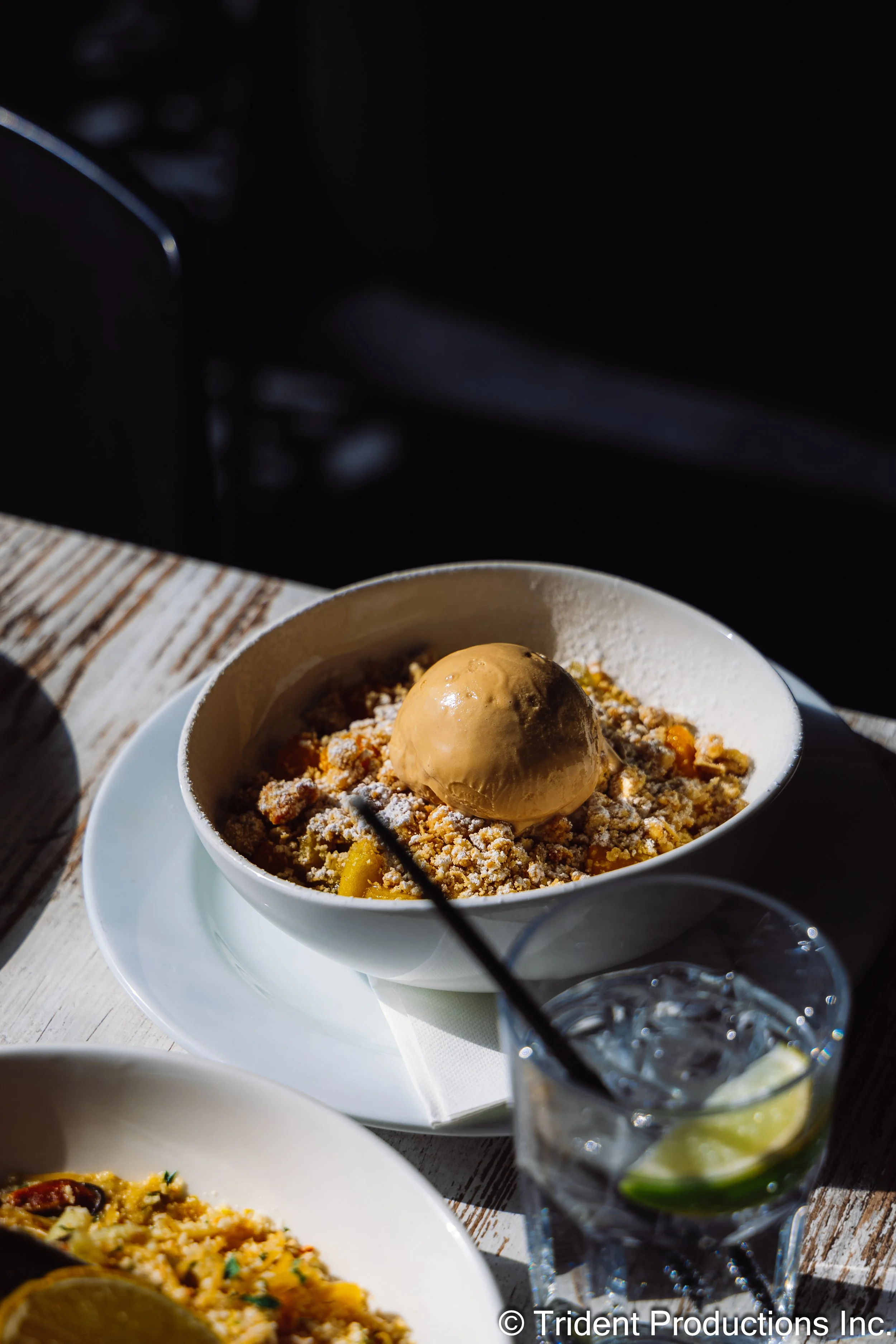 A dessert bowl with a scoop of ice cream on top, topped with powdered sugar, served with a glass of lemonade with lemon slices and ice, and a plate of food on a wooden table.