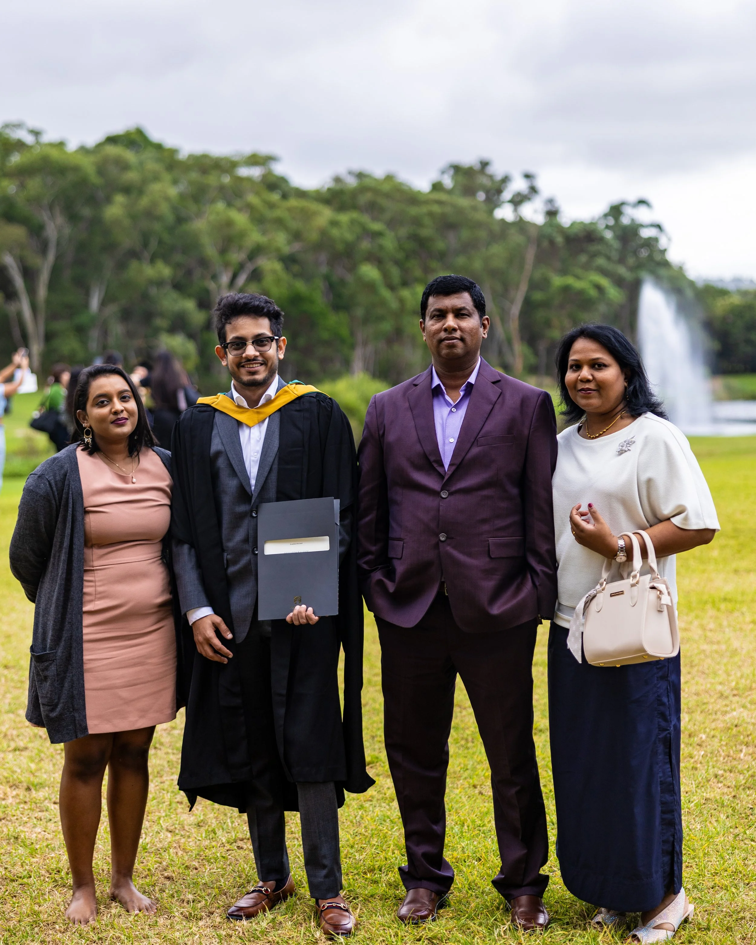 Group of four people standing on grass during daytime, celebrating a graduation, with trees and a fountain in the background.