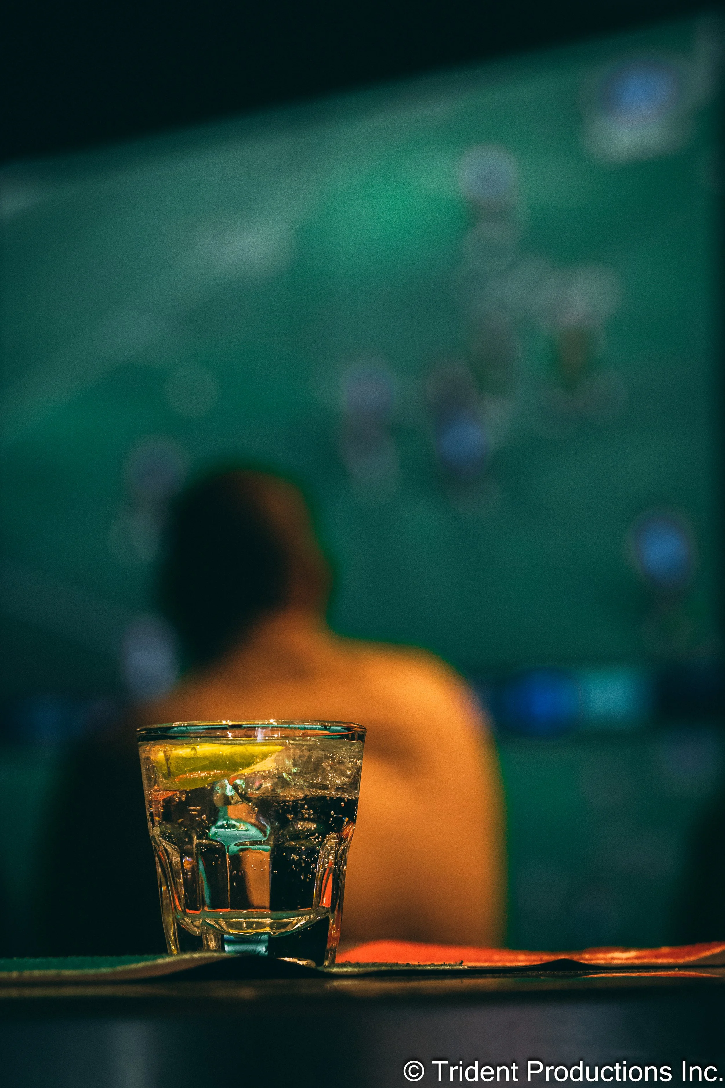 A close-up of a glass of clear liquid with ice and a lemon wedge on a table in the foreground, with a blurred person sitting in a dimly lit bar or lounge in the background.