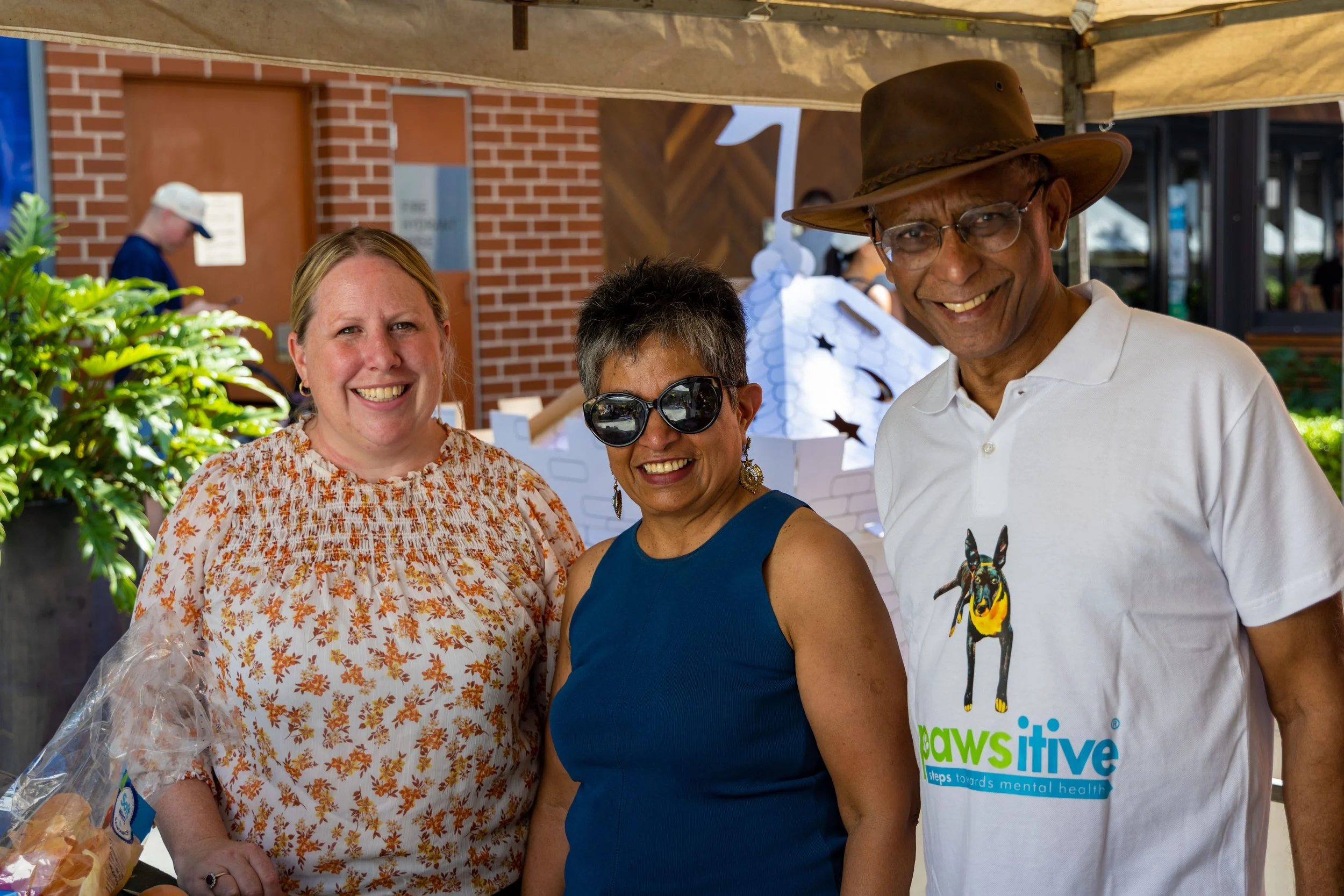 Three people smiling at an outdoor event, standing under a canopy with a brick building behind them. The woman on the left has blonde hair, is wearing a floral blouse, and has a plastic bag of baked goods on the table. The woman in the middle has sho
