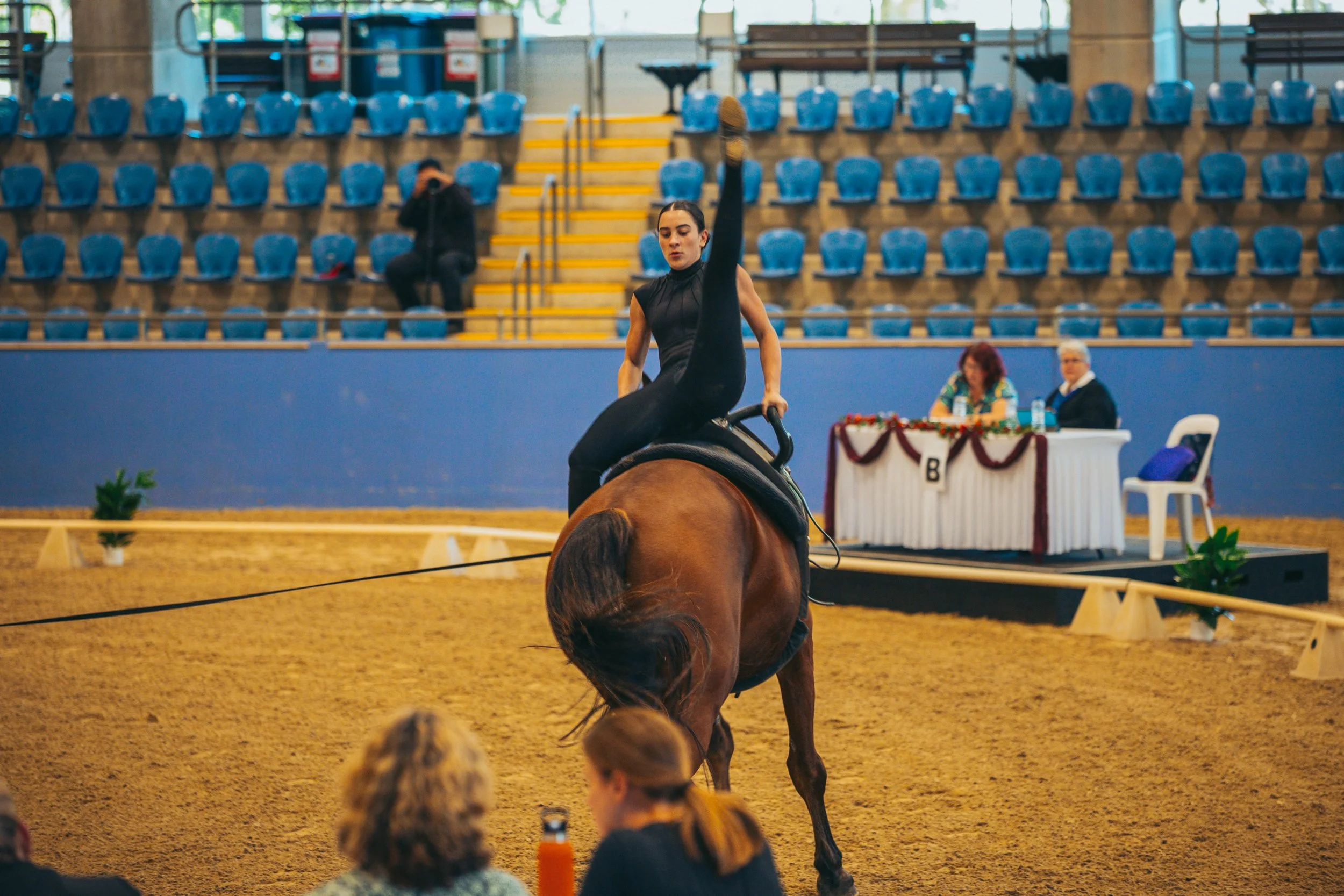An equestrian rider performs a dressage routine on a brown horse in an indoor arena. The rider is balancing on one leg with the other leg extended upward. Spectators watch in the background, with some seated and some standing. Two judges or officials