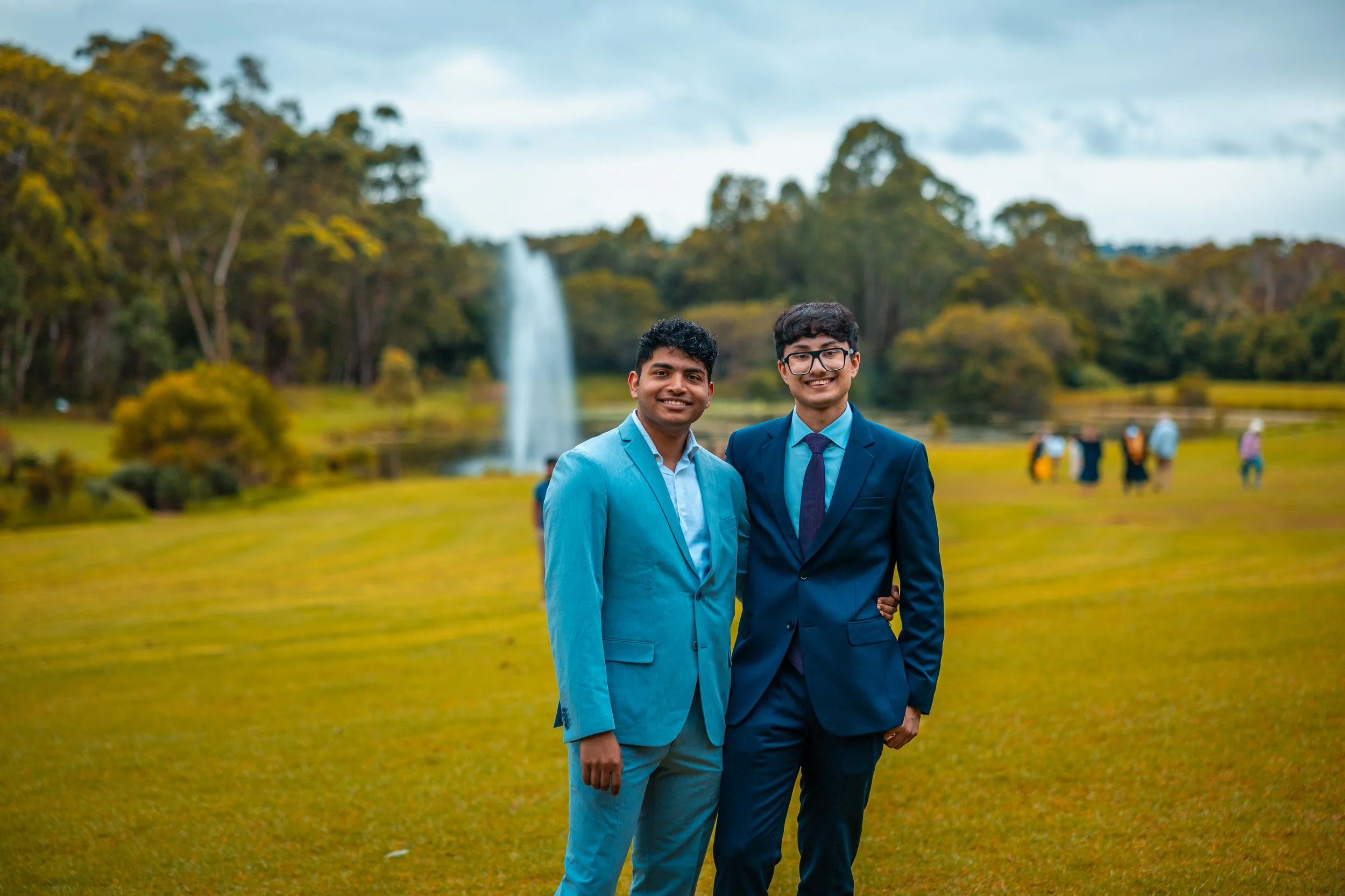 Two young men in suits standing outdoors on a grassy field, smiling, with a lake, fountain, and trees in the background.