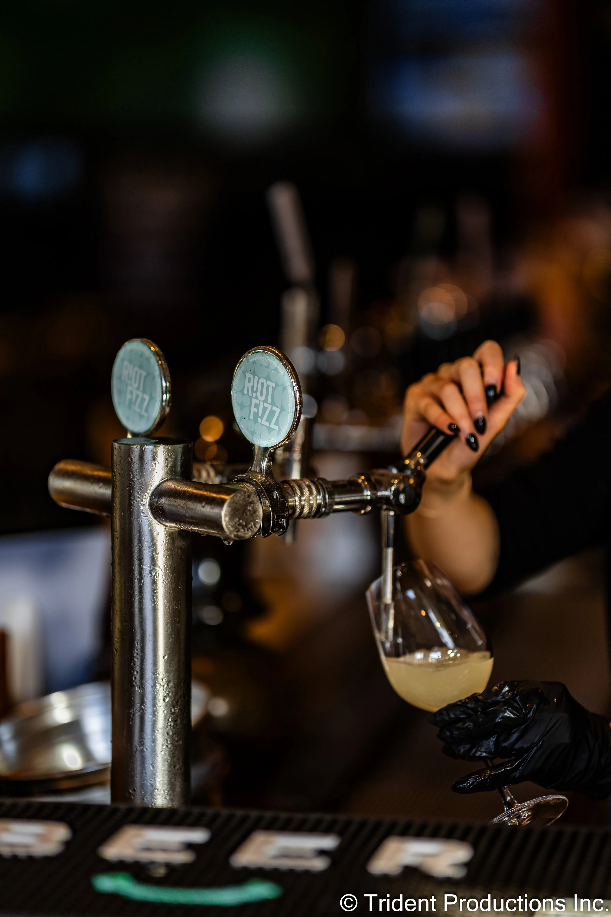 A bartender pours a drink from a tap labeled 'Riot Fizz' into a cocktail glass at a bar.
