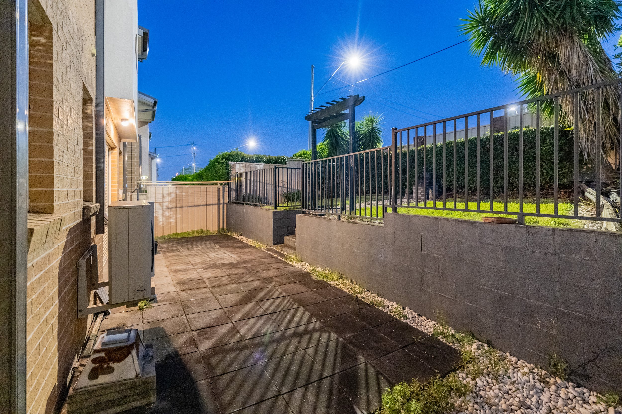 Nighttime backyard patio with brick wall, metal fence, and lush greenery.