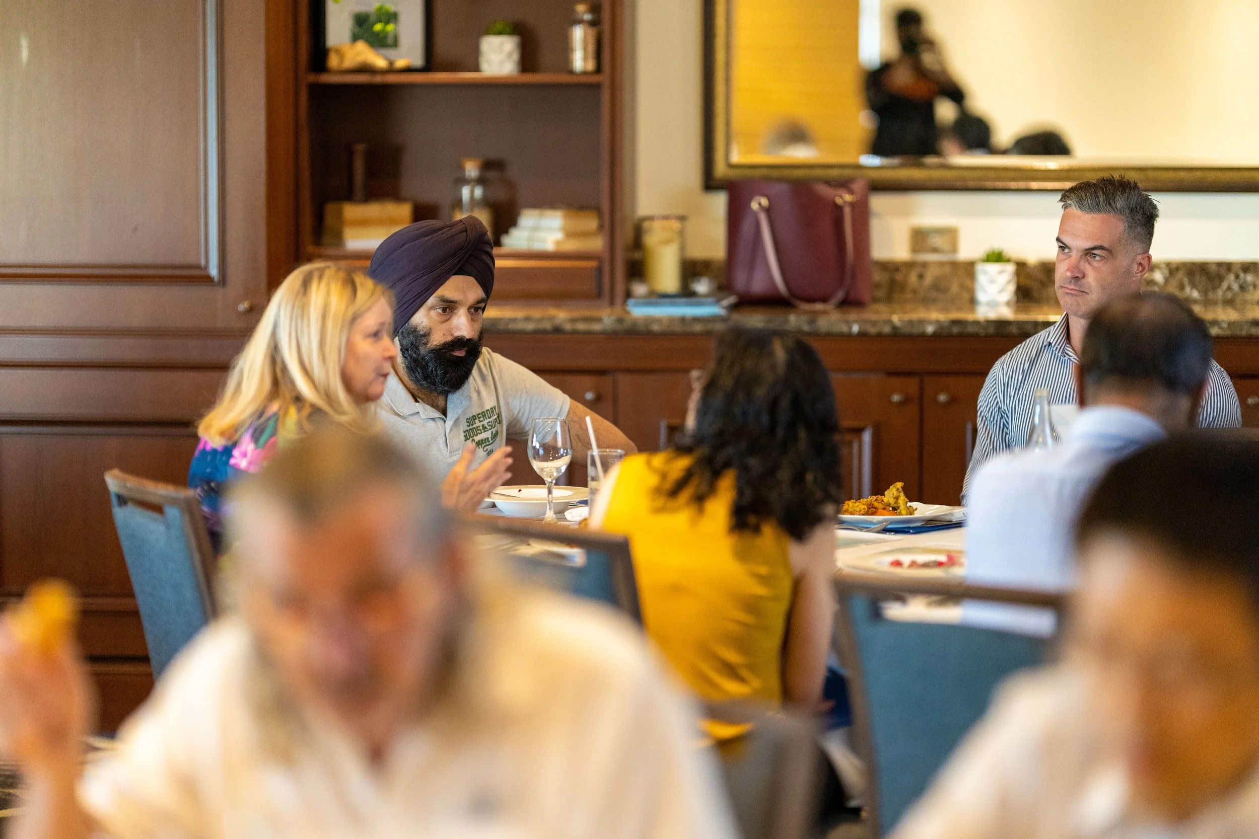Group of people sitting at a dinner table in a restaurant, engaged in conversation. There is a woman with blonde hair, a man with a turban and beard, and a man with gray hair. The background shows a wooden cabinet, a mirror, and a granite countertop.