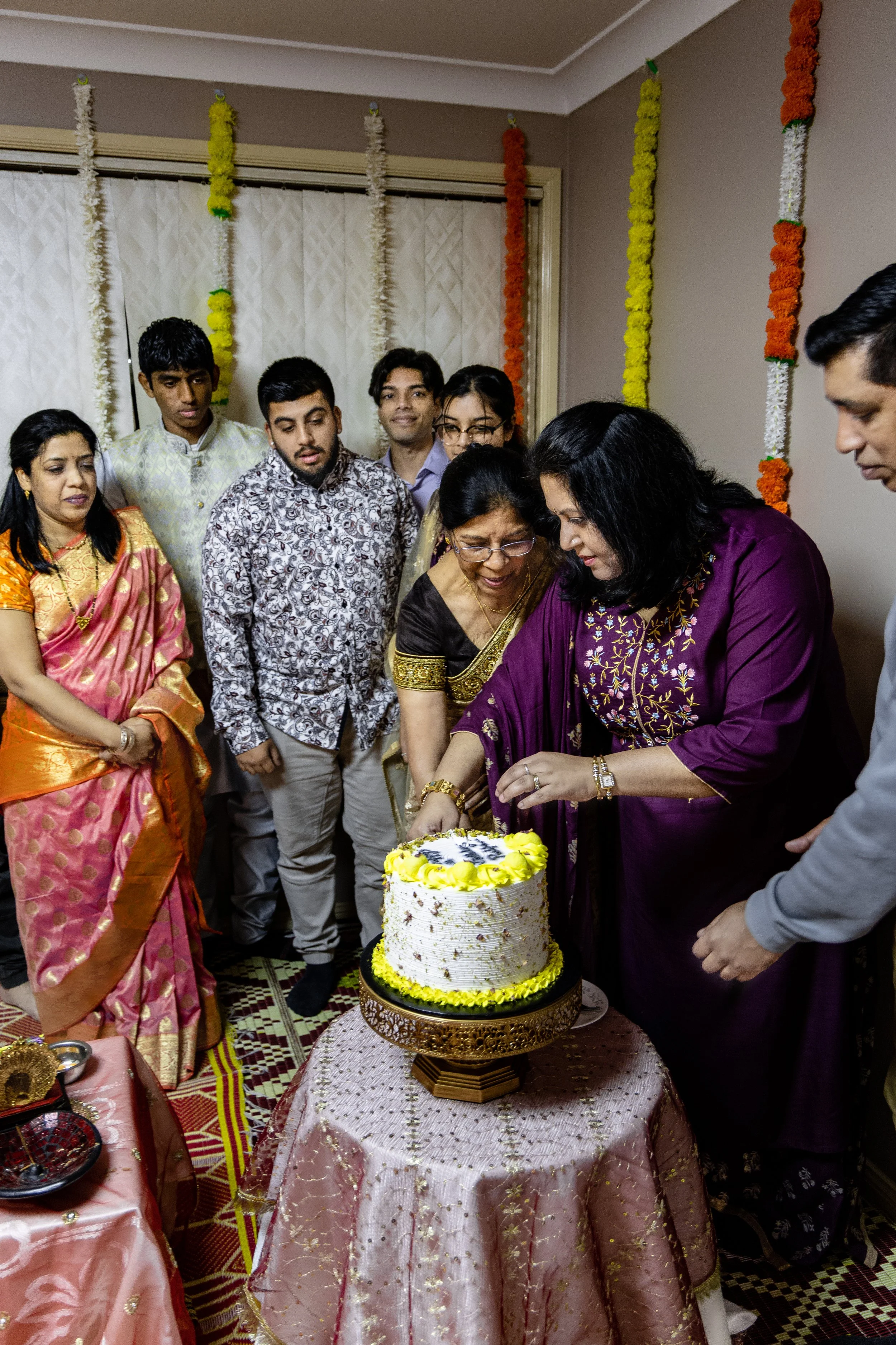 Group of Indian people celebrating a birthday with a cake, with women in traditional sarees and men in traditional ethnic clothing, gathered around a decorated table with colorful marigold garlands hanging in the background.